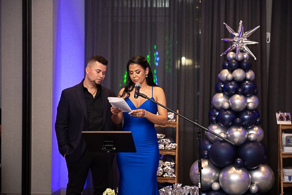 Man and woman at event, woman in blue dress reading from notes, microphone, balloon tree.