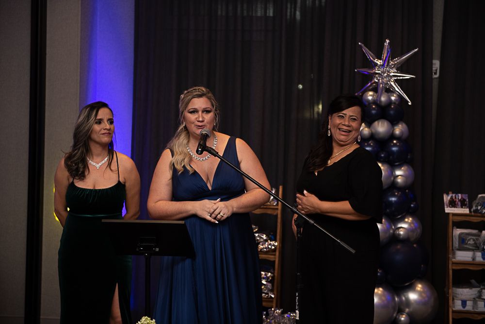 Three women in evening gowns speaking at an event with blue and silver decorations.