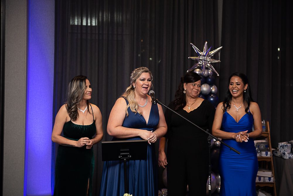 Four women in evening gowns on a stage. One speaks into a microphone, others smile, a balloon display in the background.