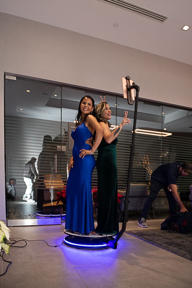 Two women in formal dresses pose on a photo booth platform, smiling and pointing. Modern interior.