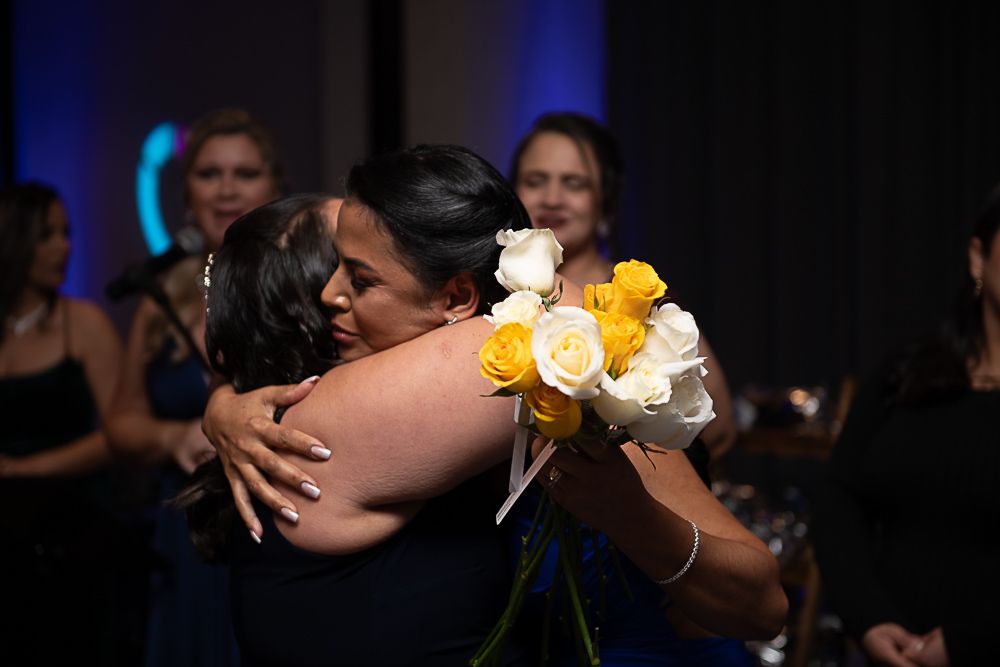 Two women embrace, one holding yellow and white roses. Other women watch in a dark setting.