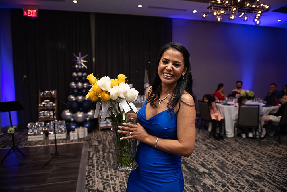 Woman in blue dress holding flowers, smiling at event with decorations and guests.