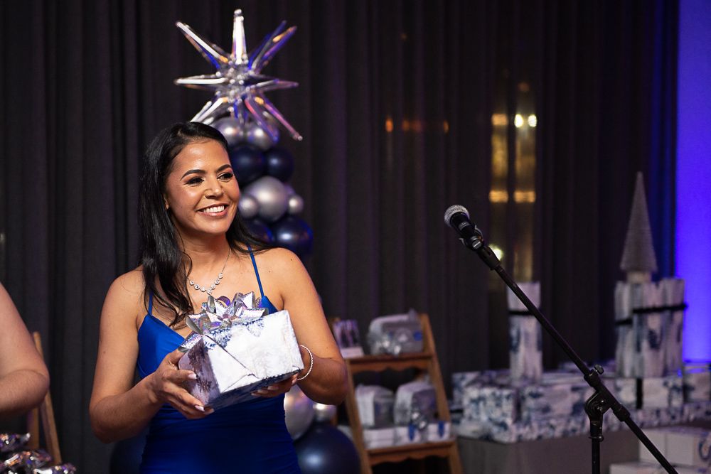 Woman in blue dress holding a gift, smiling at an event. Dark setting with balloons and mic.