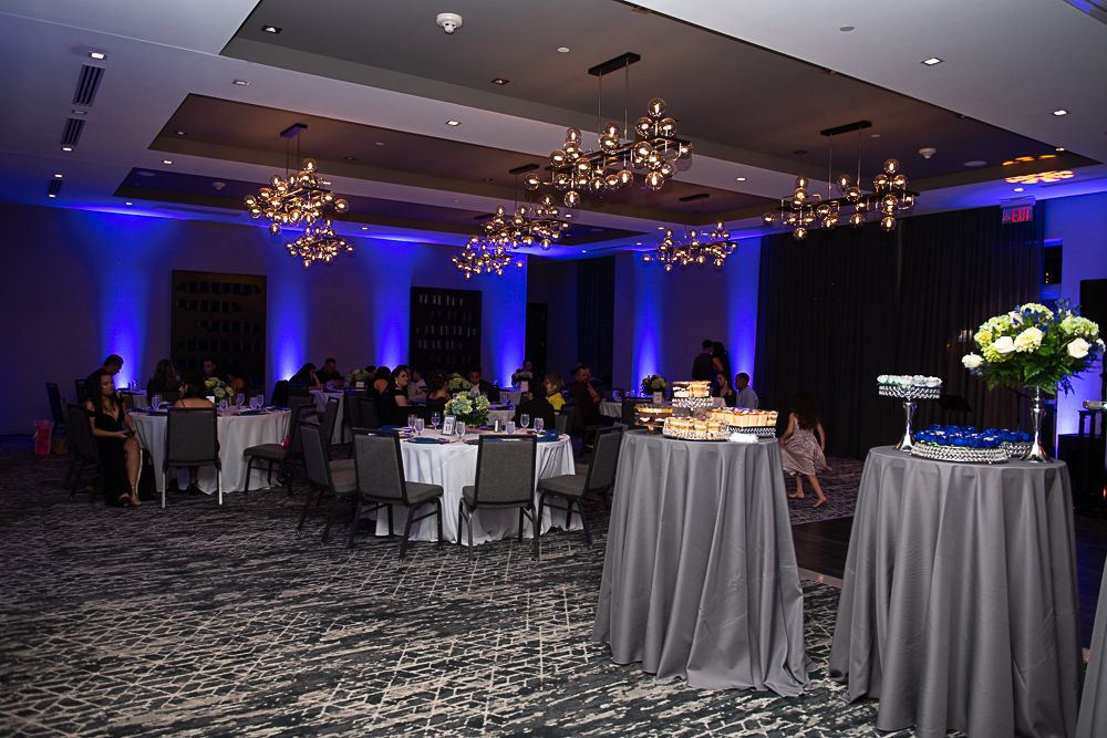 Reception hall with tables, guests, blue lighting, and chandeliers.