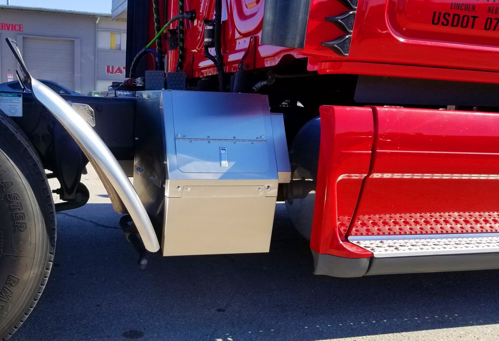 Close-up of a red truck cab with a silver fuel tank and black step on a sunny street.