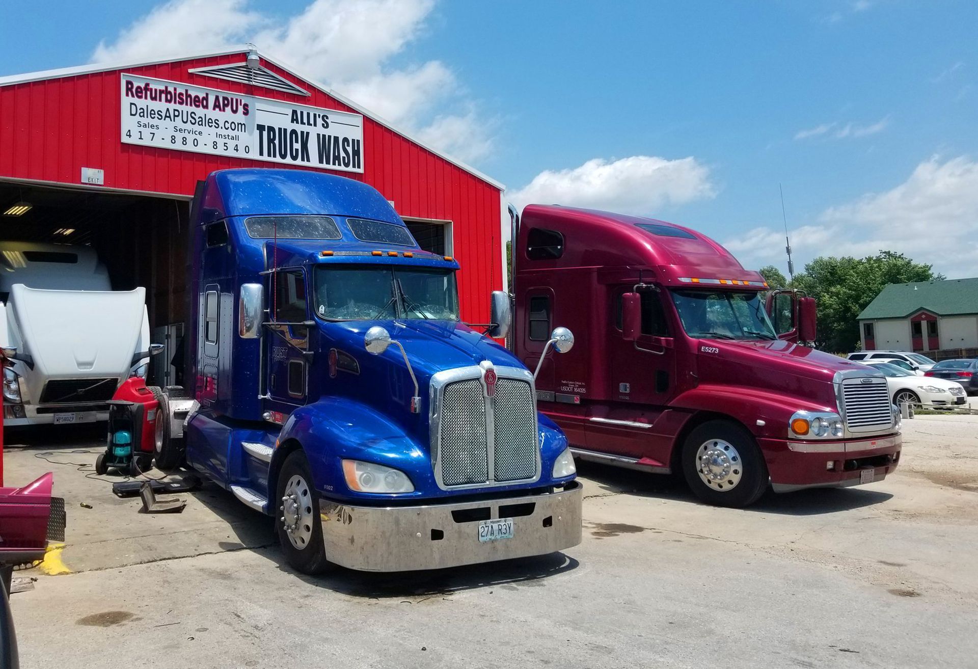 Two semi trucks, blue and red, parked outside a truck repair shop under a blue sky