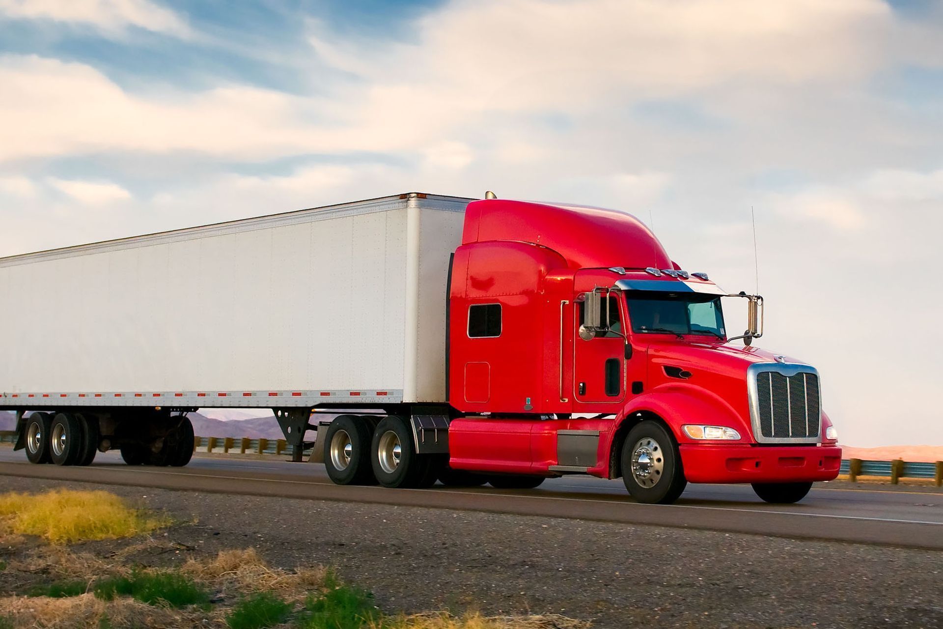 Red semi-truck hauling a white trailer on a highway under a cloudy sky
