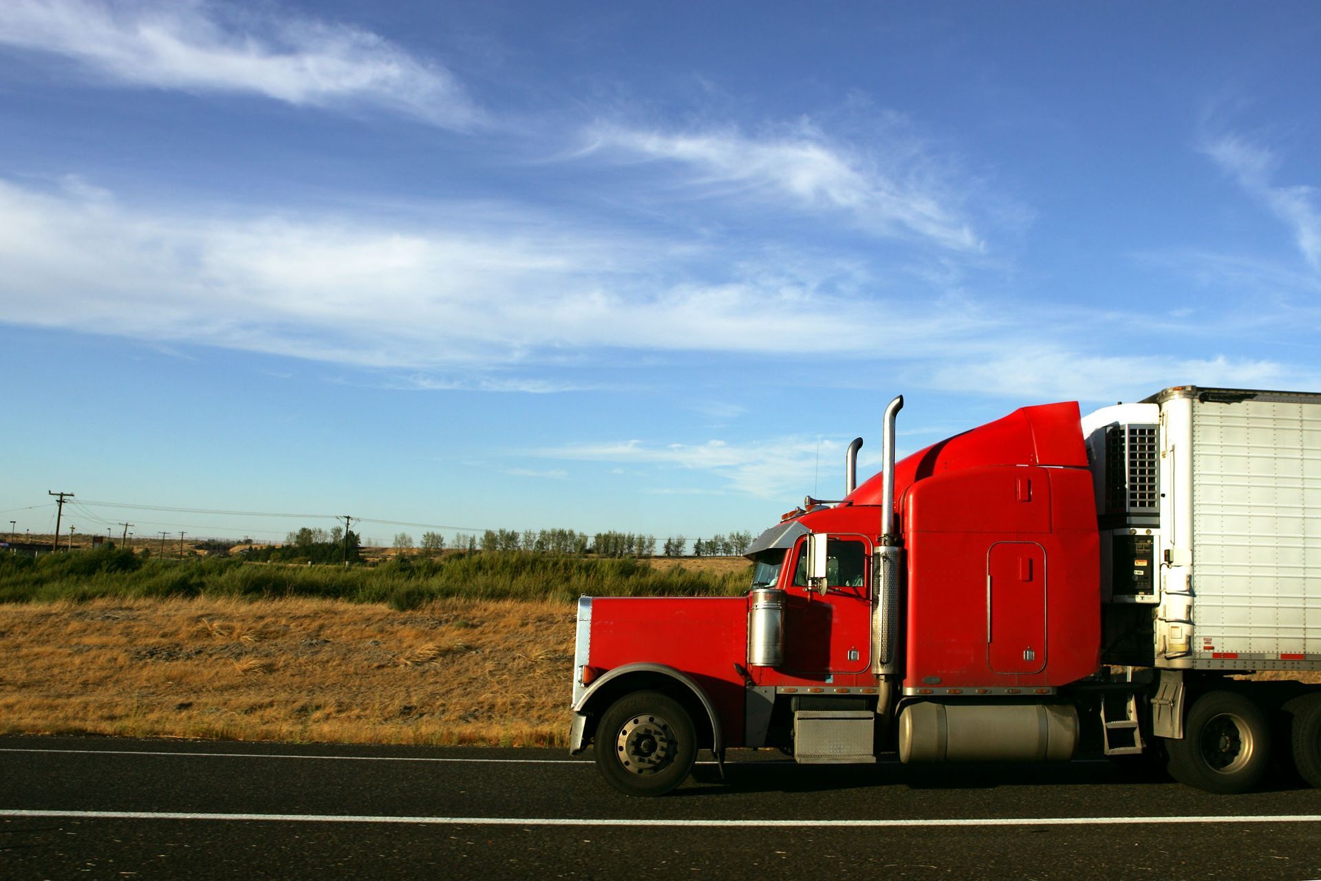 A red semi-truck driving along a highway next to an open field under a blue, cloudy sky.