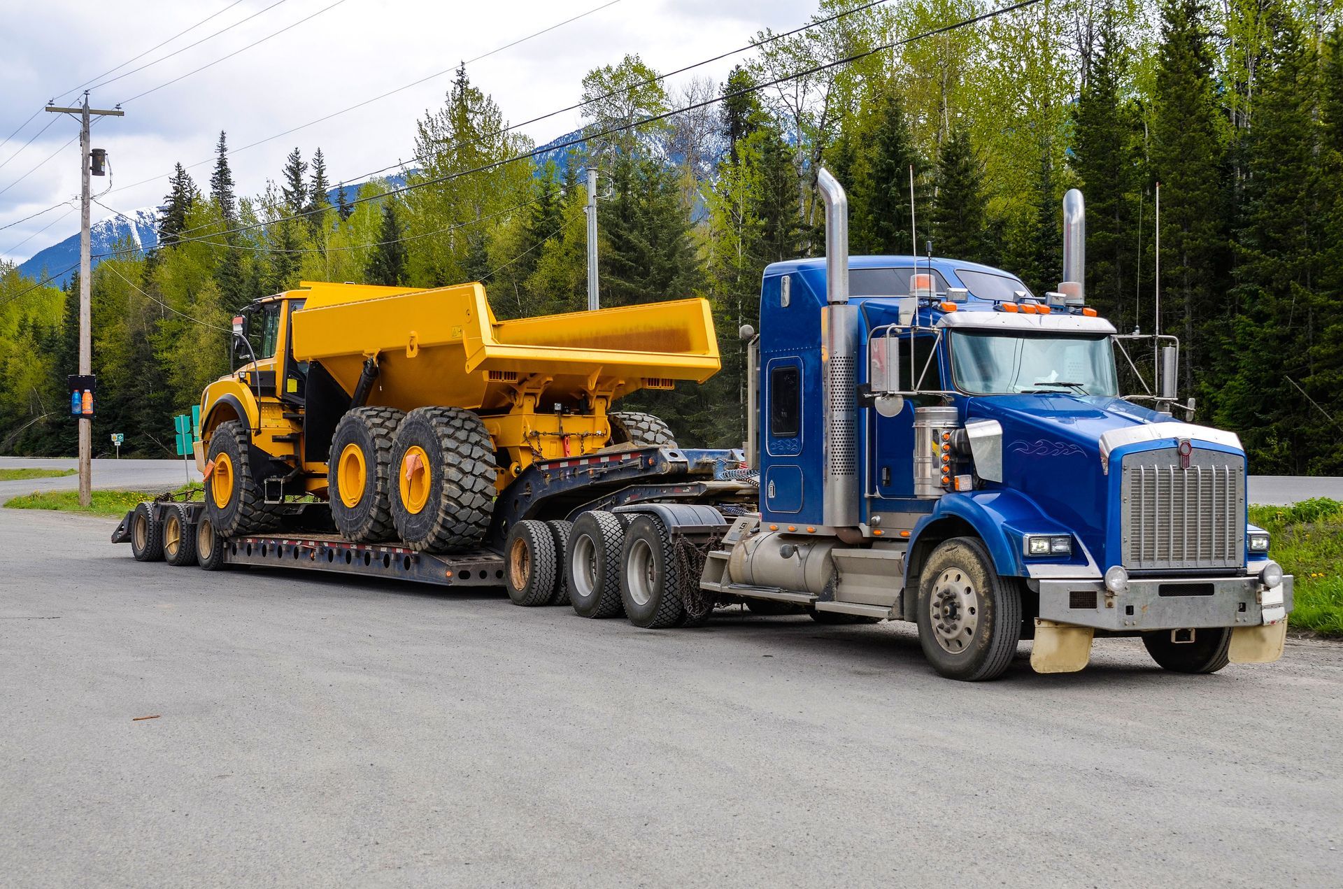 Blue semi truck hauling a yellow dump truck on a lowboy trailer beside a forested road