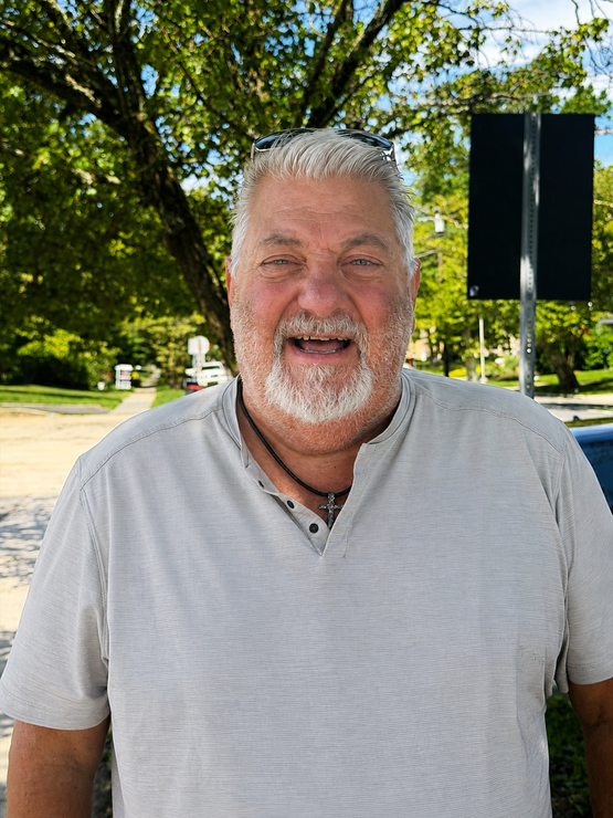 Smiling man in a light gray shirt standing outdoors on a sunny day.