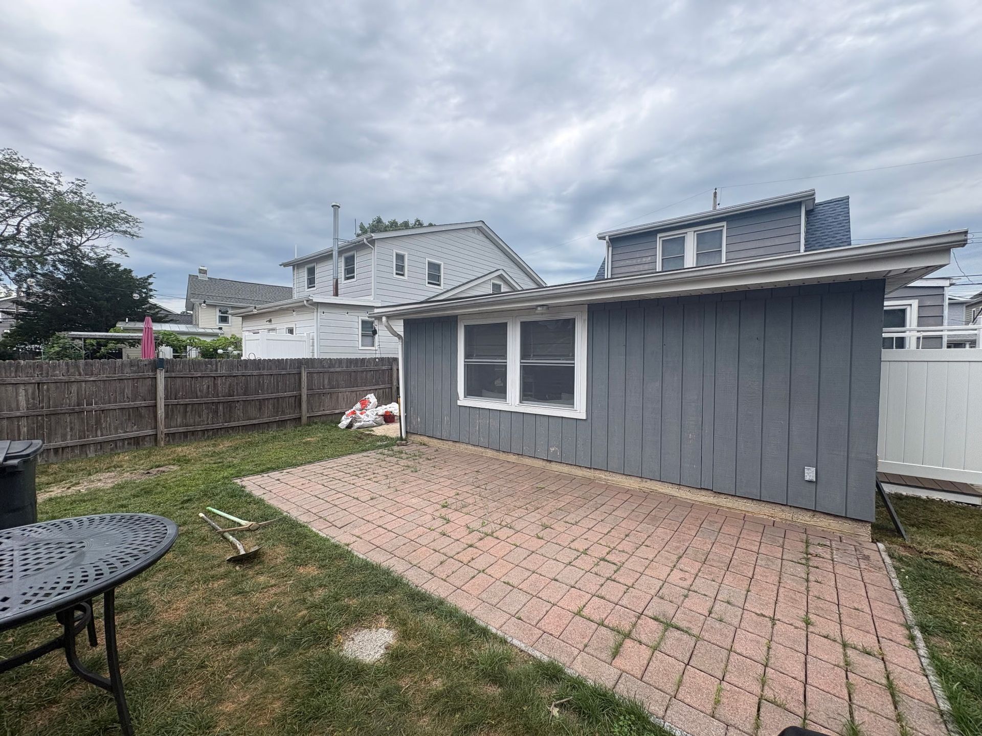 Backyard patio with gray shed, brick pavers, grass, fence, and cloudy sky