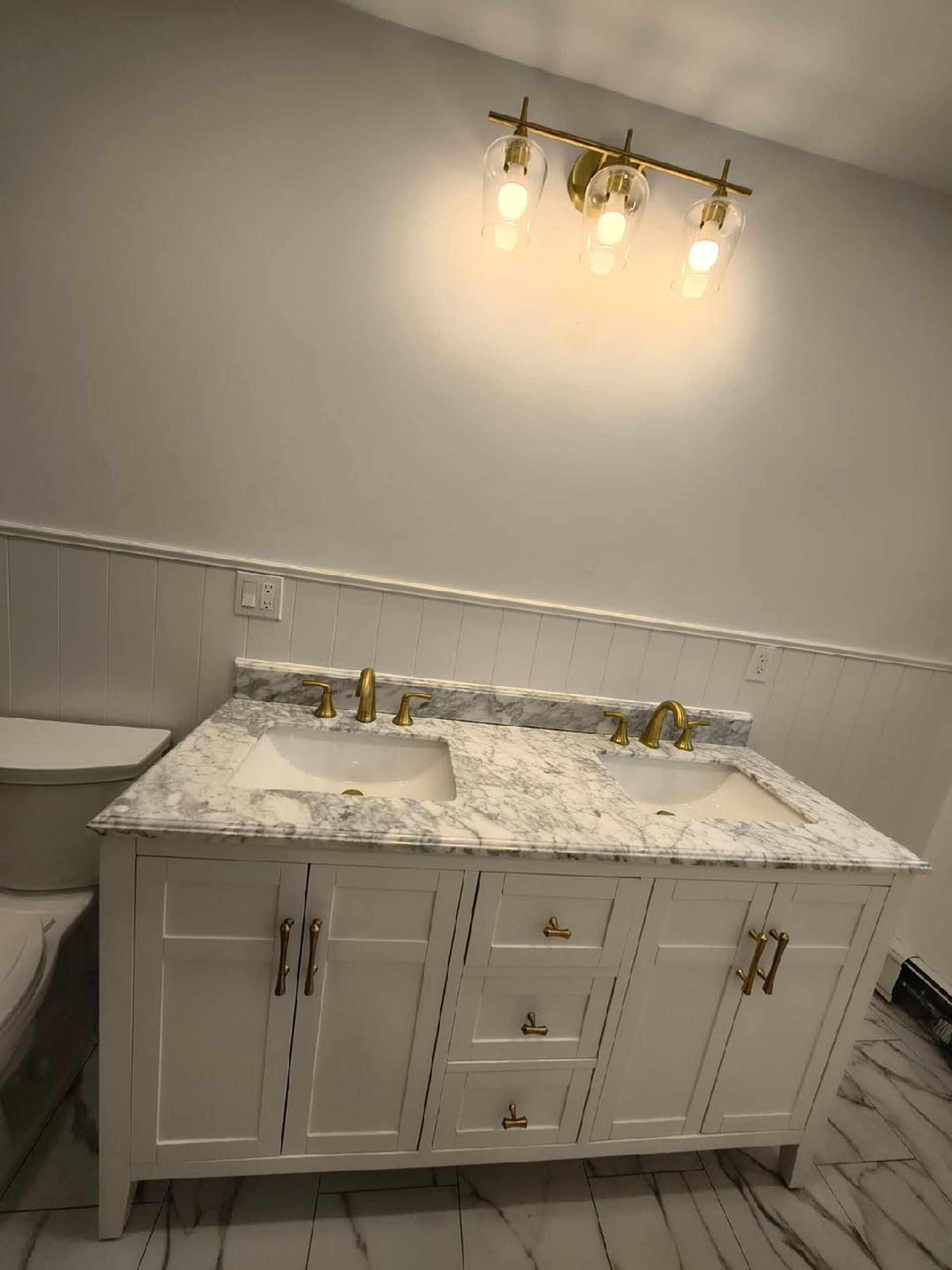 White double vanity with marble countertop and gold fixtures, under a four-light vanity fixture.