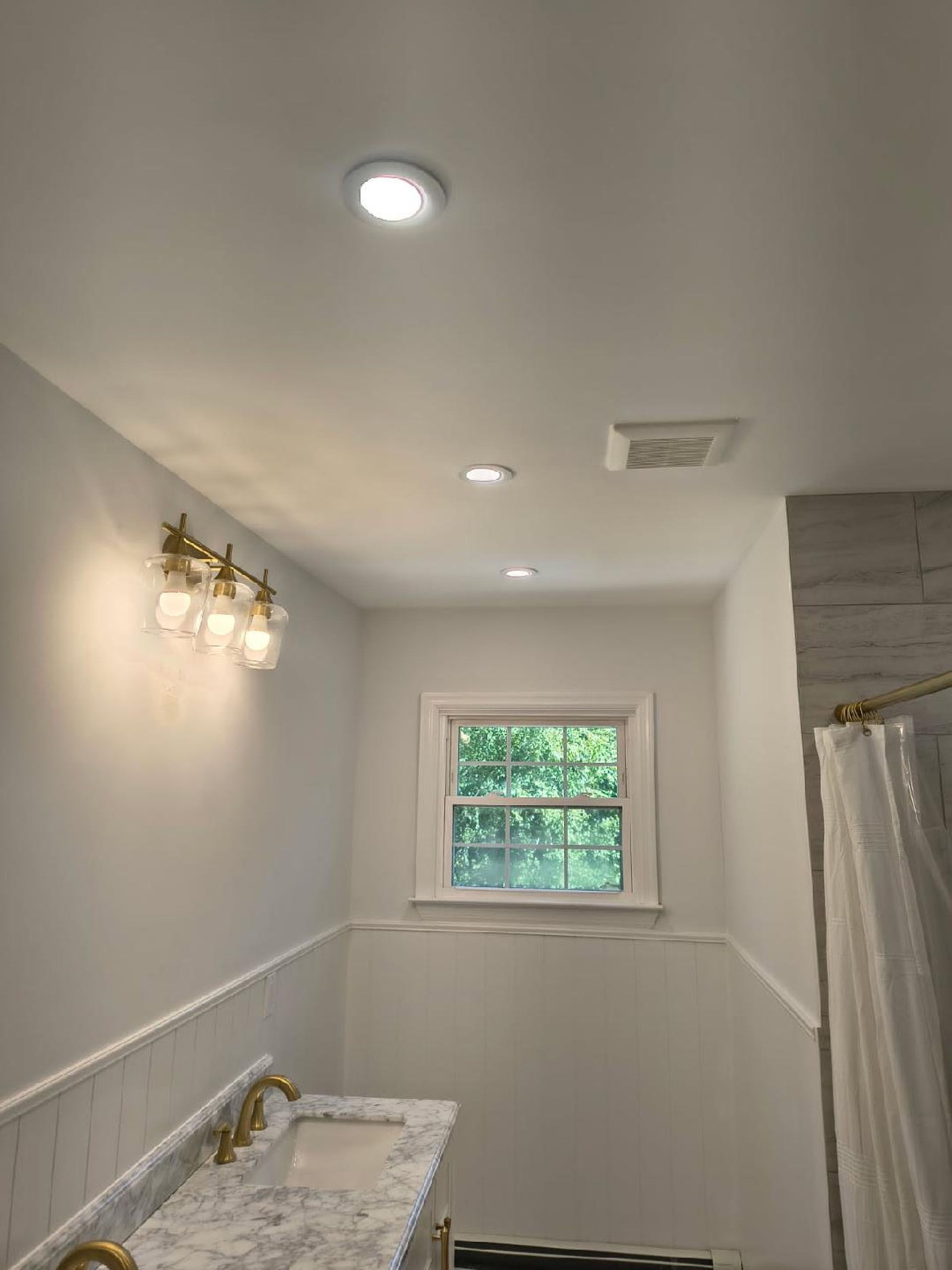 Bathroom interior with overhead lighting, a window, vanity with gold fixtures, and a shower.