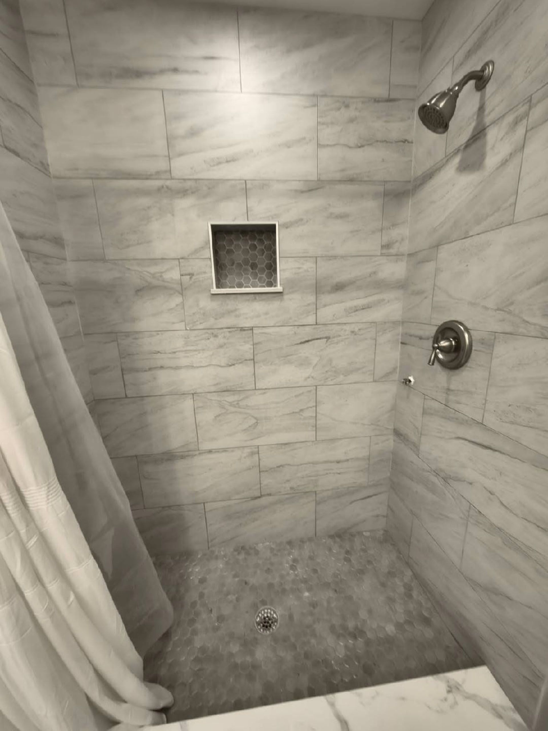 Shower with marble-like tiles and a built-in shelf. A white curtain hangs on the left.