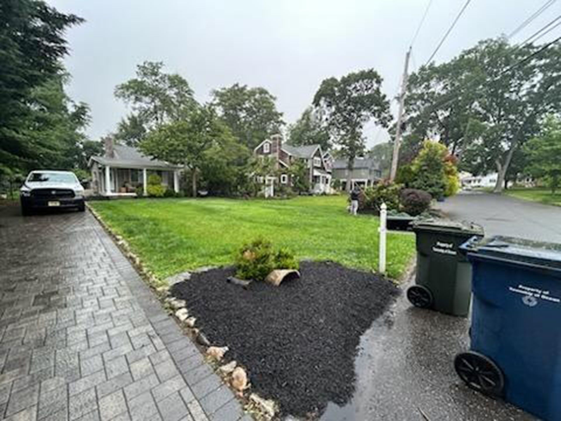 Lawn and homes on a residential street. A truck is parked on a paved driveway. Trash cans are in the right foreground.