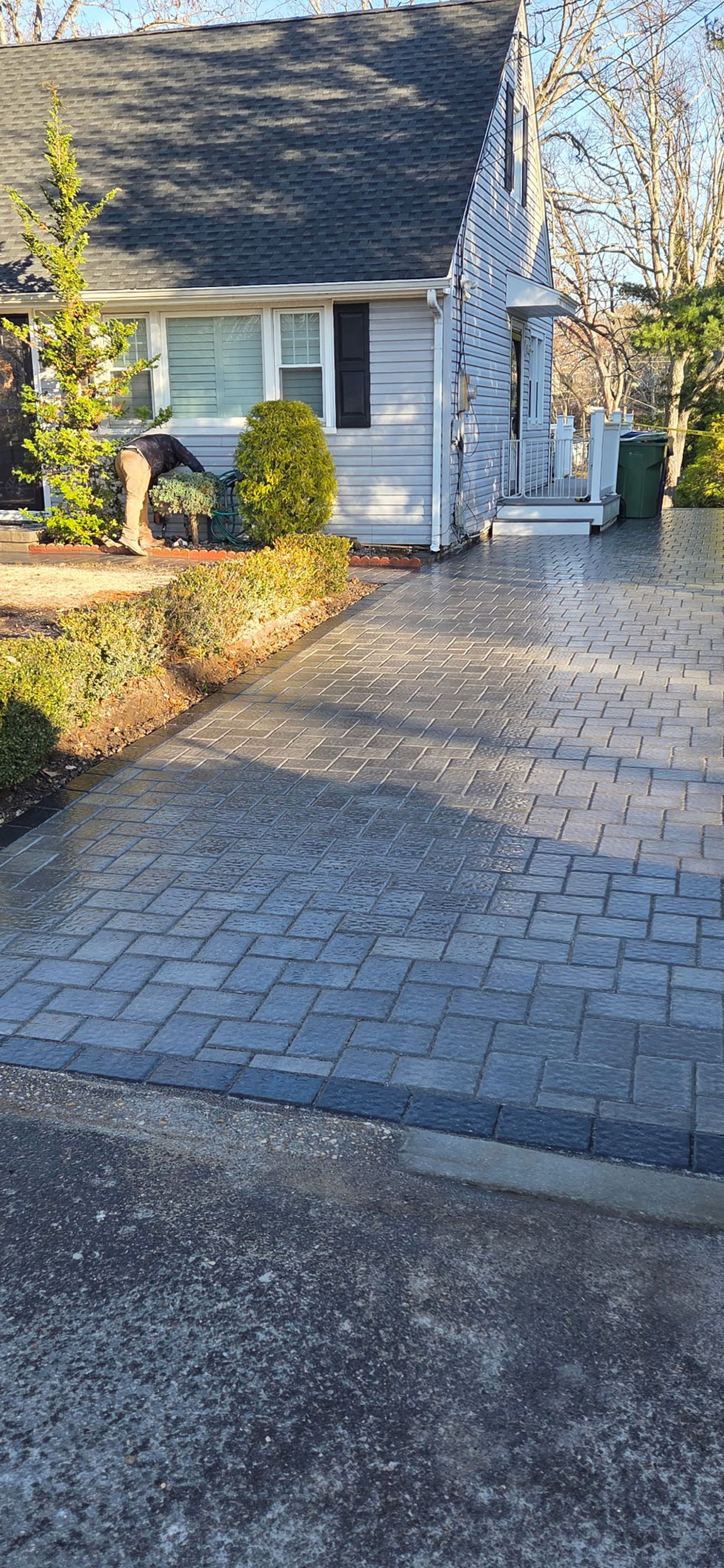 A gray house with a dark roof and a brick driveway. Green bushes and a small tree flank the driveway.
