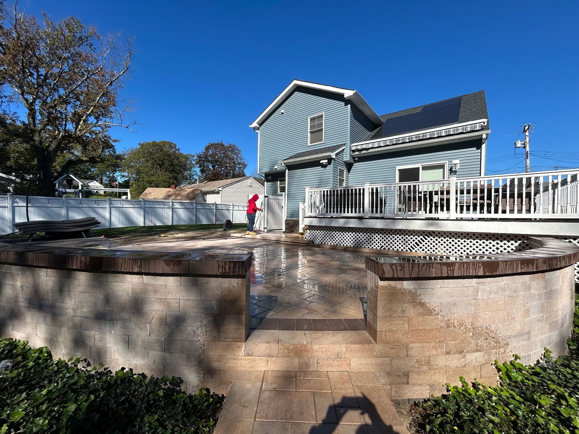 Backyard with brick patio, wooden deck, and blue house under clear sky. Person spraying surface.