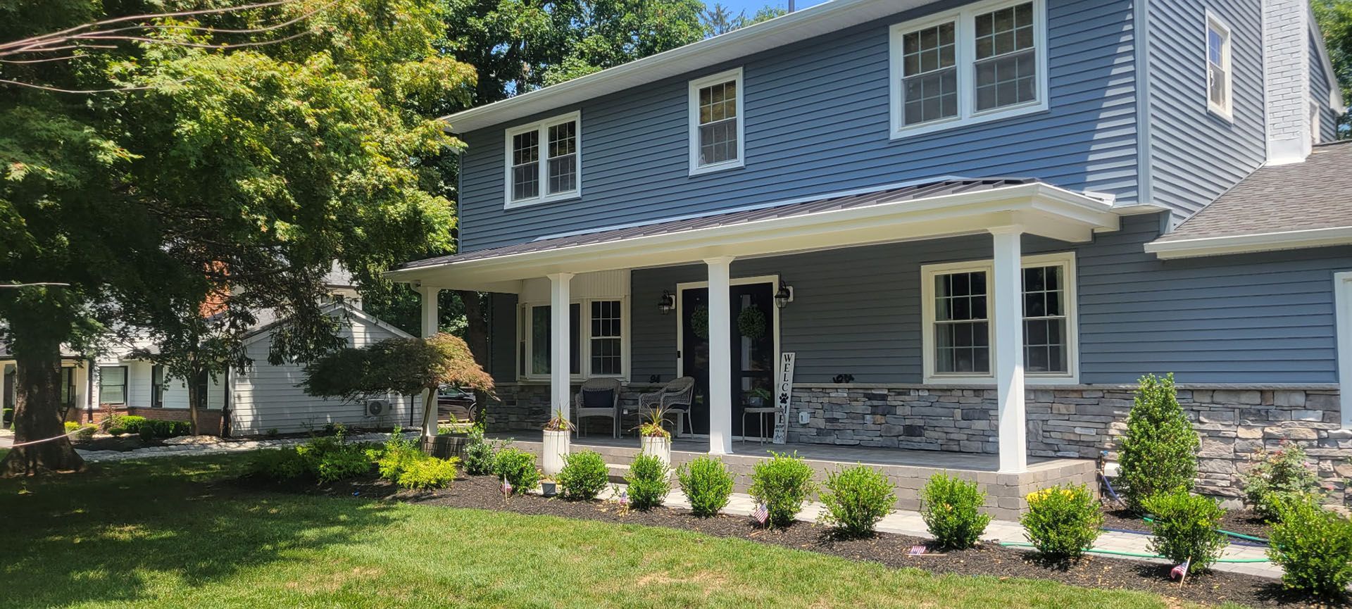 Blue-sided two-story house with a porch. Landscaping includes green bushes and lawn.