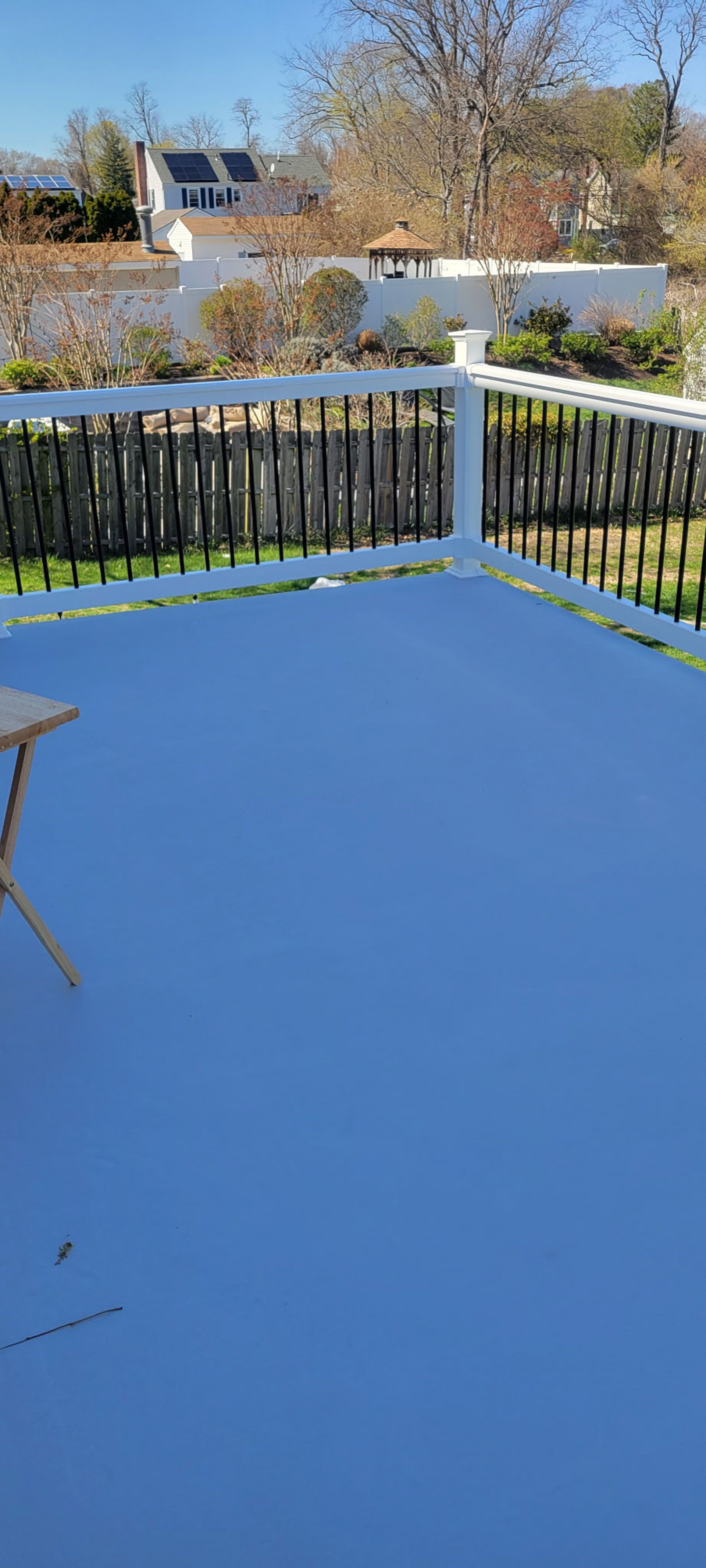 Snow-covered deck with a black railing, overlooking a yard and houses on a sunny day.