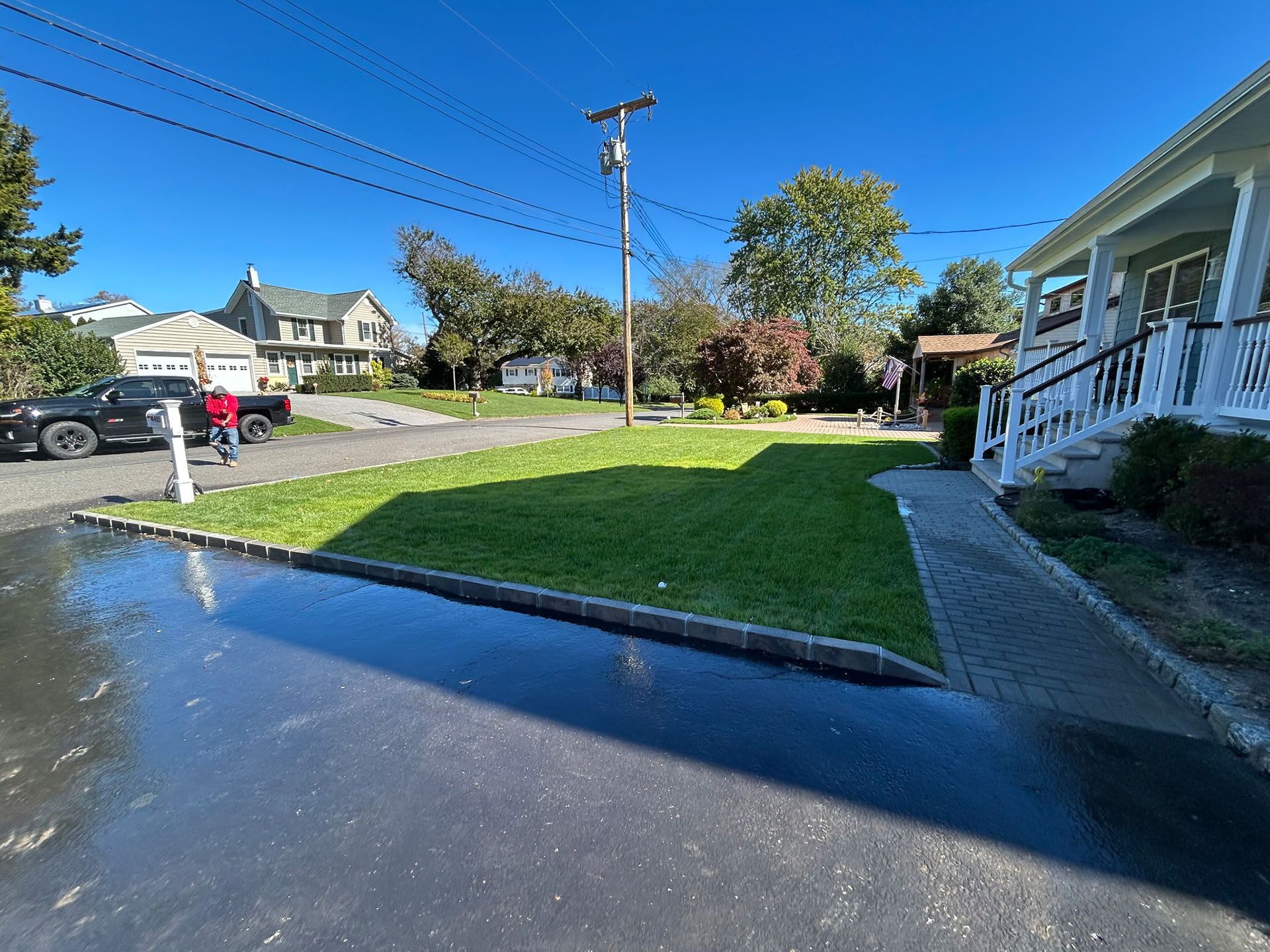 Lawn next to paved road, bordered by a stone edge, with a house on the right, and trees in the background.