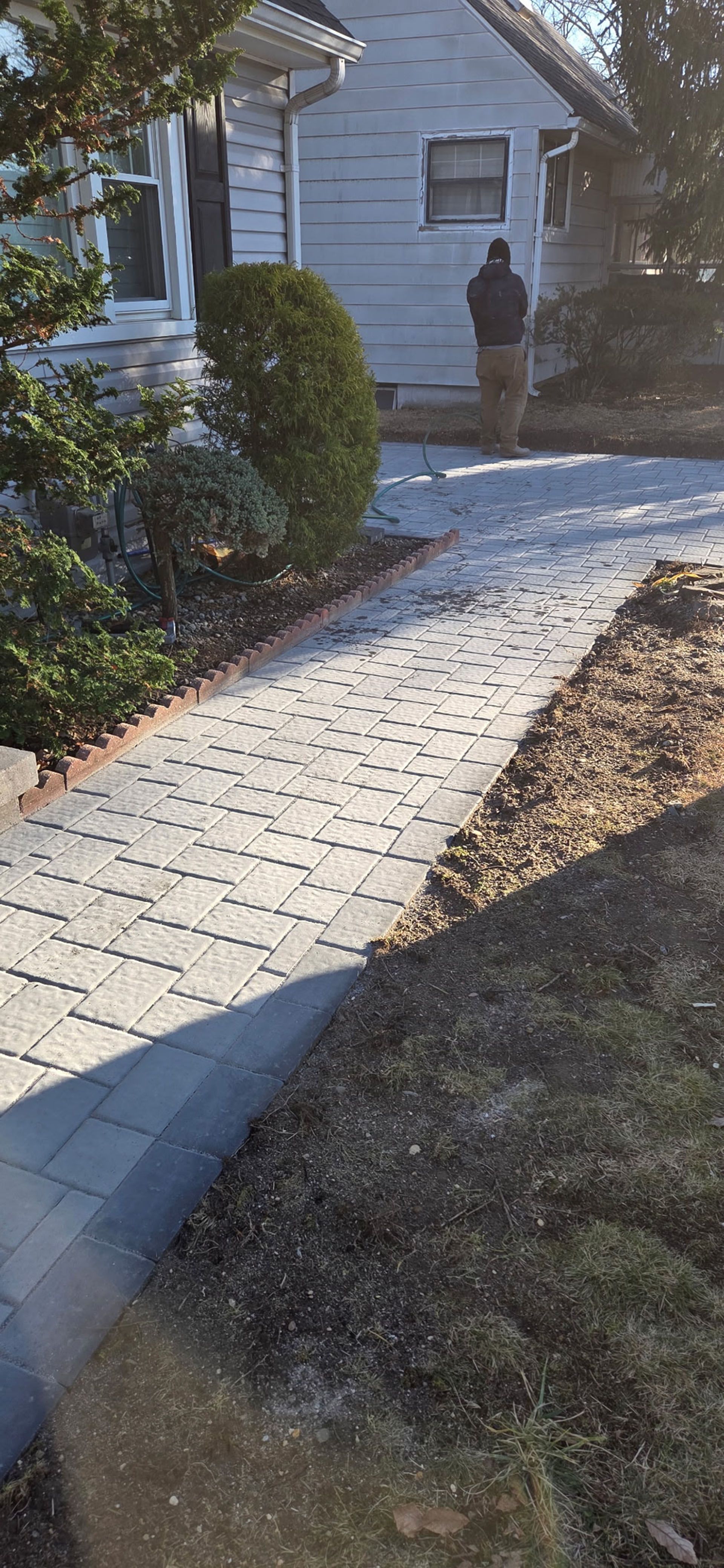 A person stands in front of a house, along a stone walkway. Shrubs and a tree are visible.