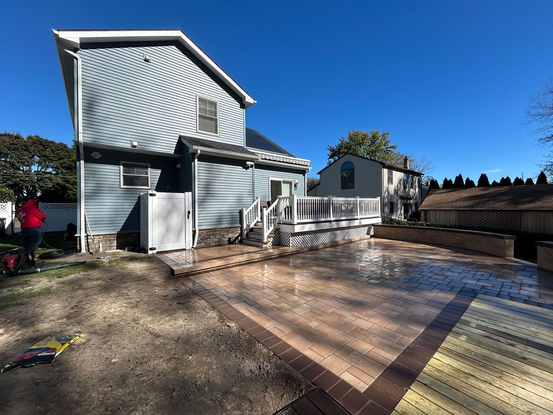 Backyard with house and new paved patio. Blue siding, wooden deck, brown brick border, clear blue sky.