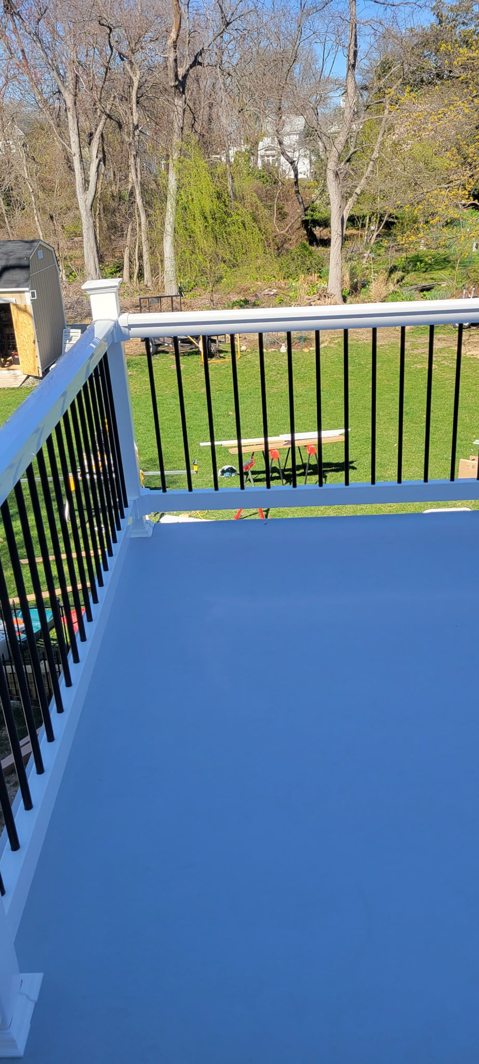 A deck with black railings overlooks a grassy yard and trees, under a blue sky.