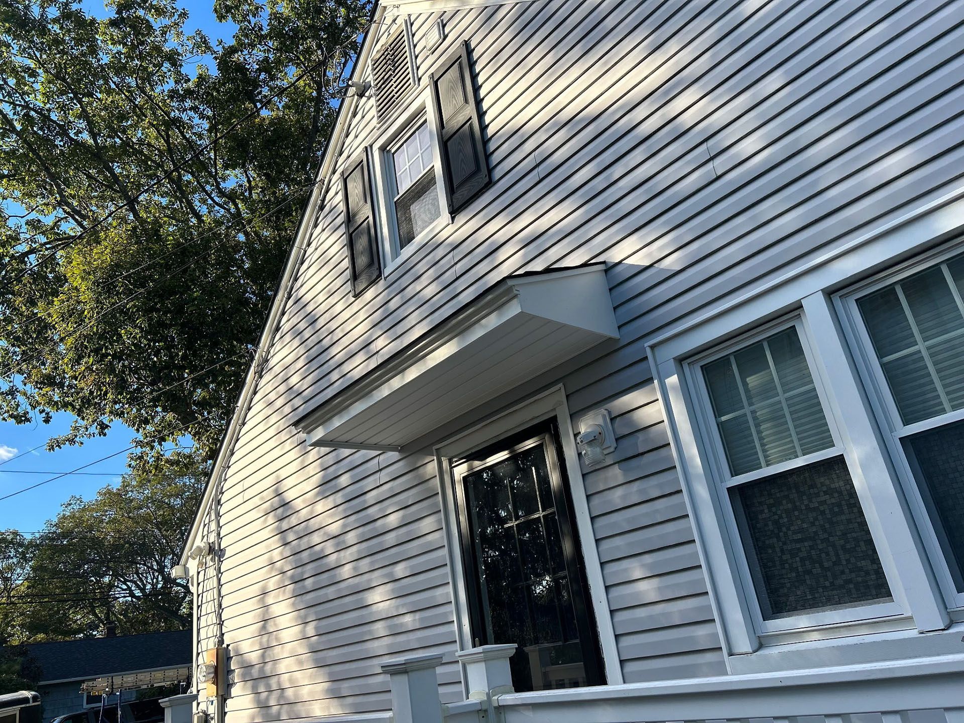 Light gray house with black front door, shutters, and an awning above the entrance. Sunny day.