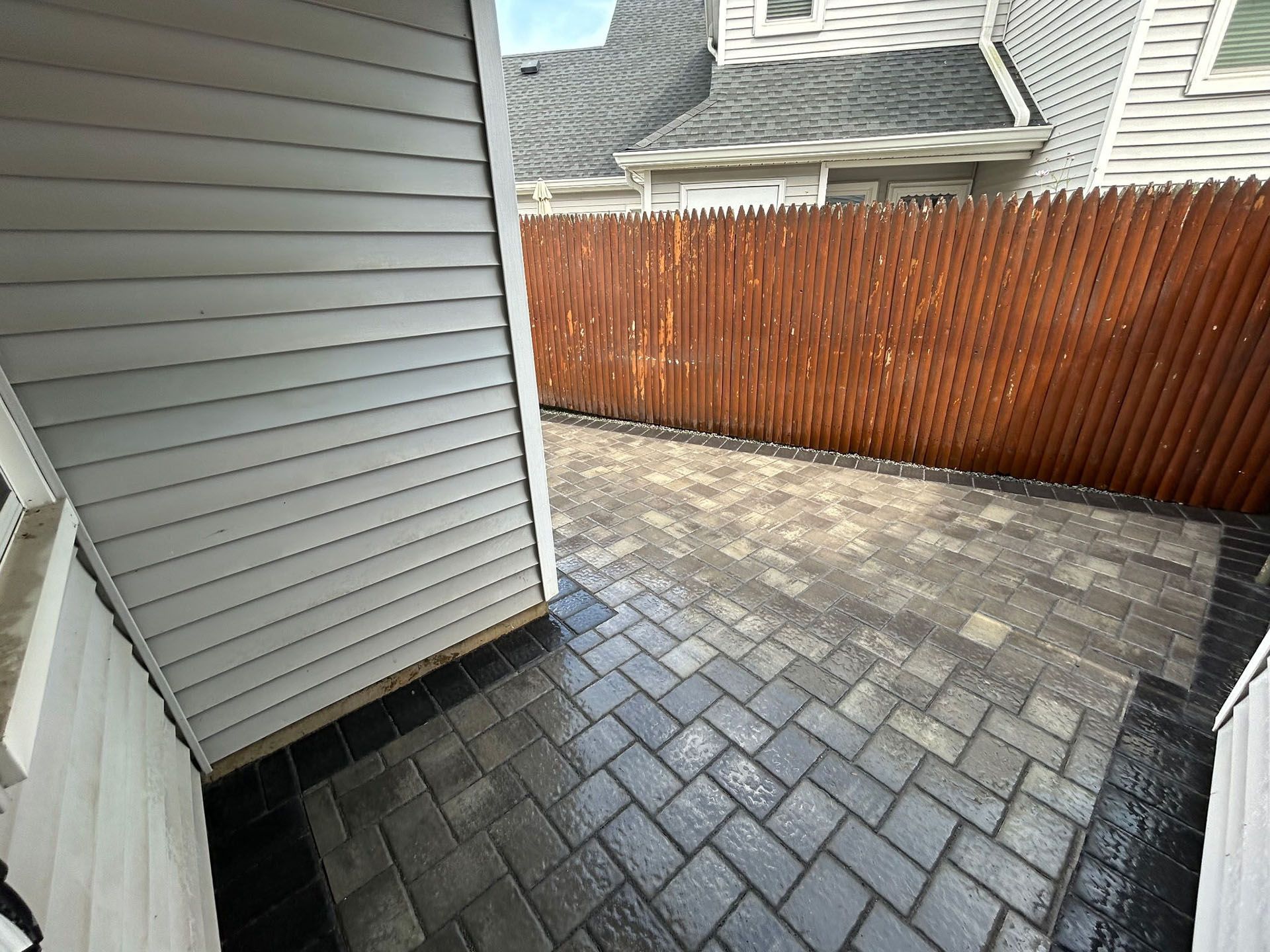 Paver patio next to a light-colored building and a wooden fence.