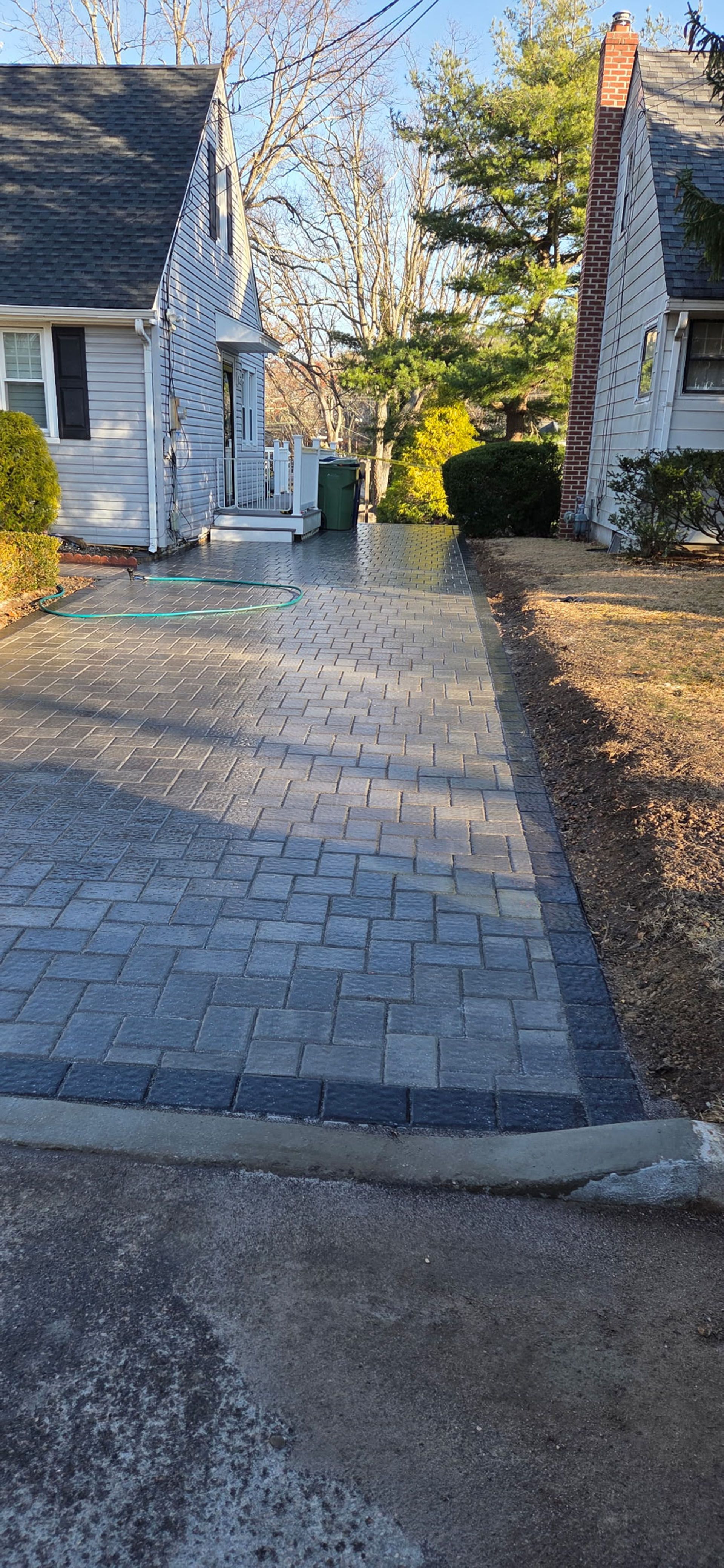 Driveway paved with gray bricks between two houses with green foliage in the background.