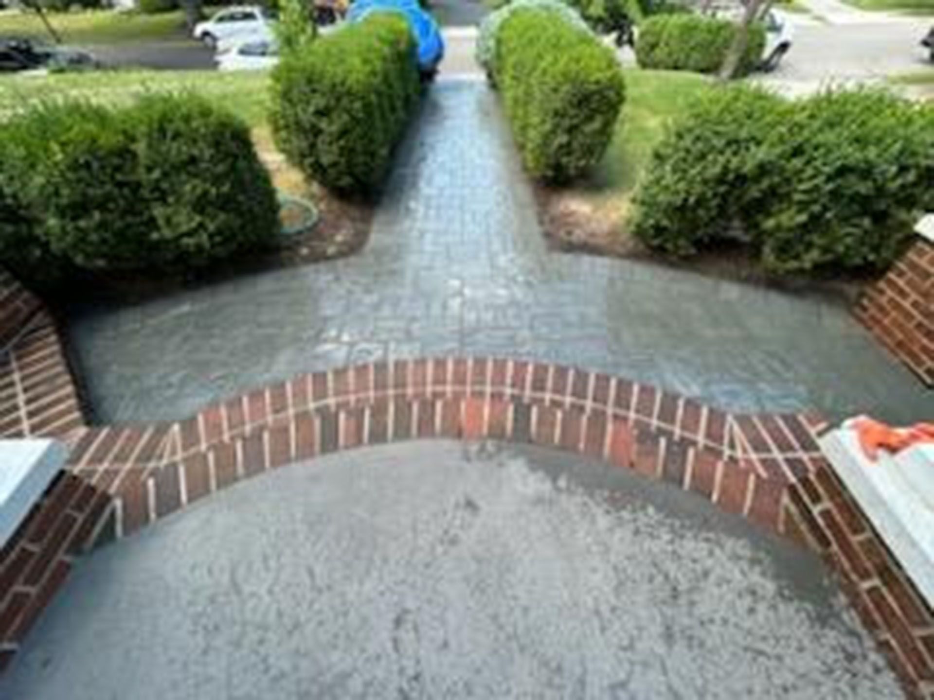 Brick arched entrance to a gray brick path lined with green bushes, leading to a street with cars.
