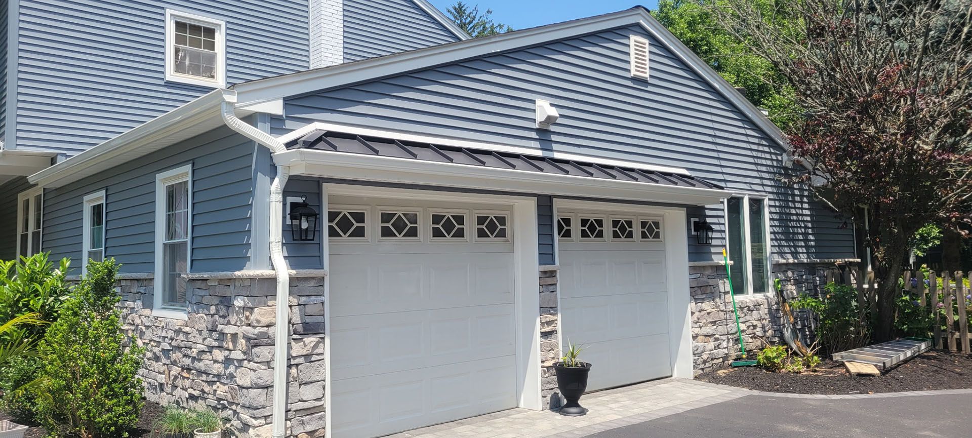 Blue house with white garage doors and stone accents, surrounded by greenery.