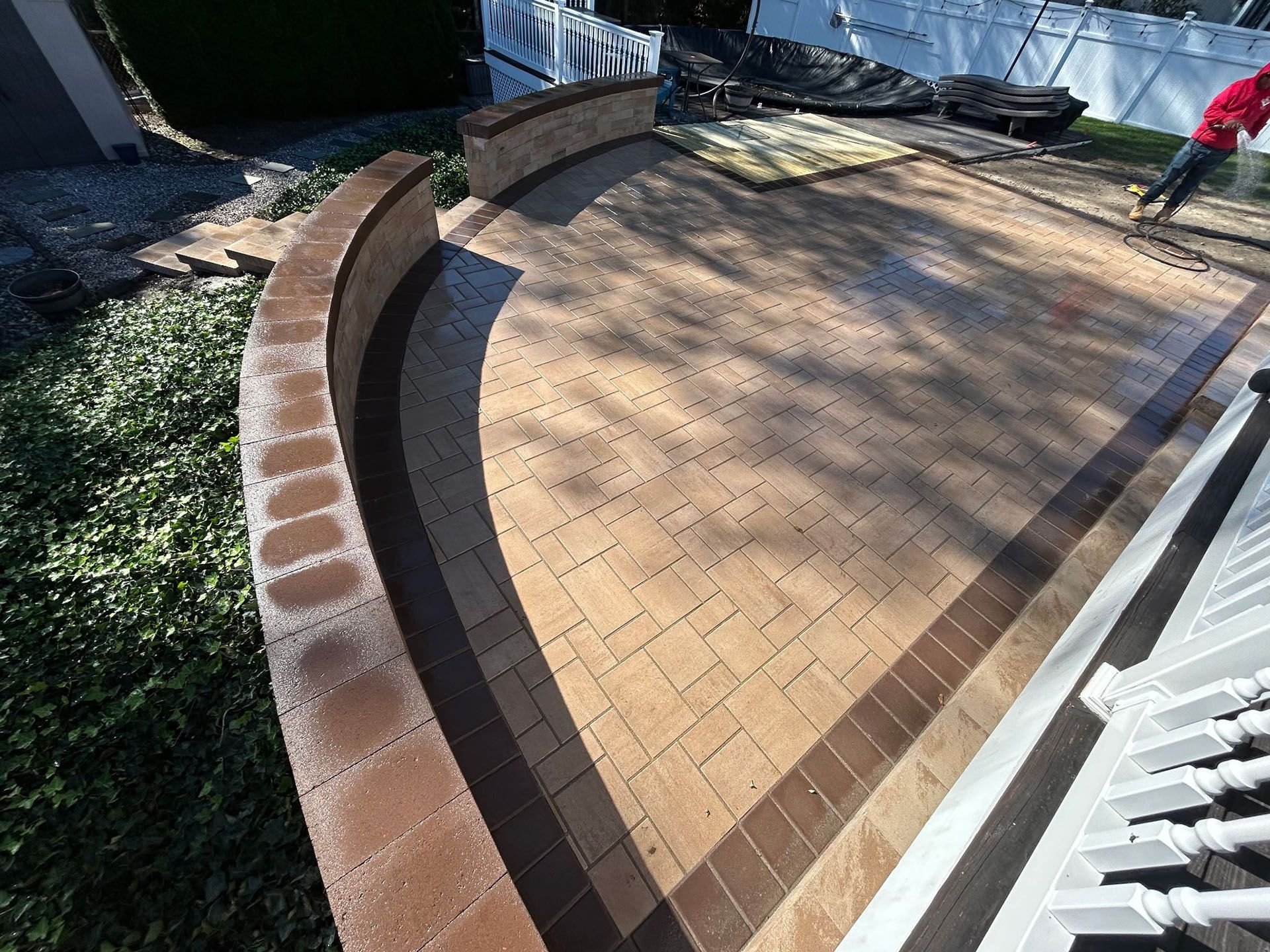 Brick patio with curved steps and retaining wall. Brown and tan pavers, white railing, greenery.