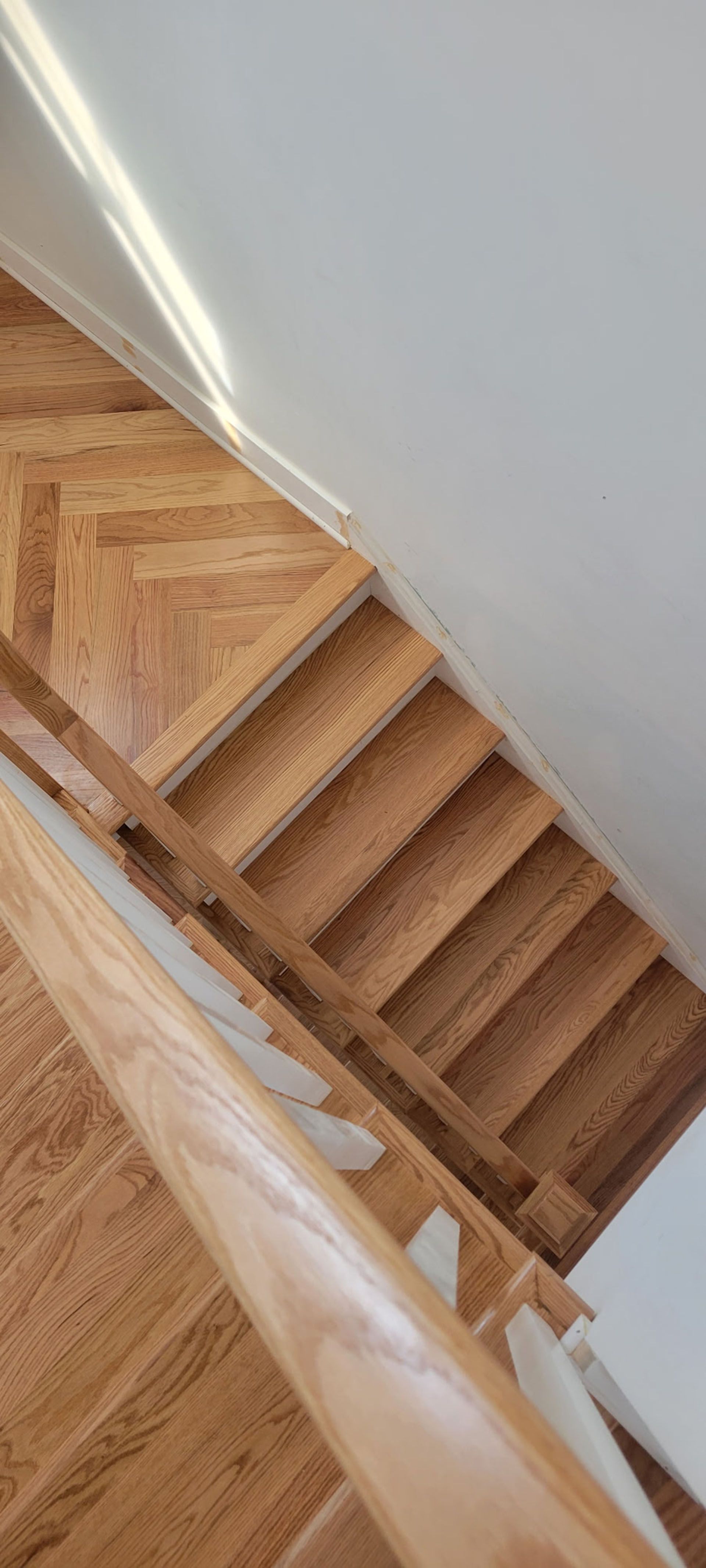 Wooden staircase with a handrail, leading upwards, against a white wall.