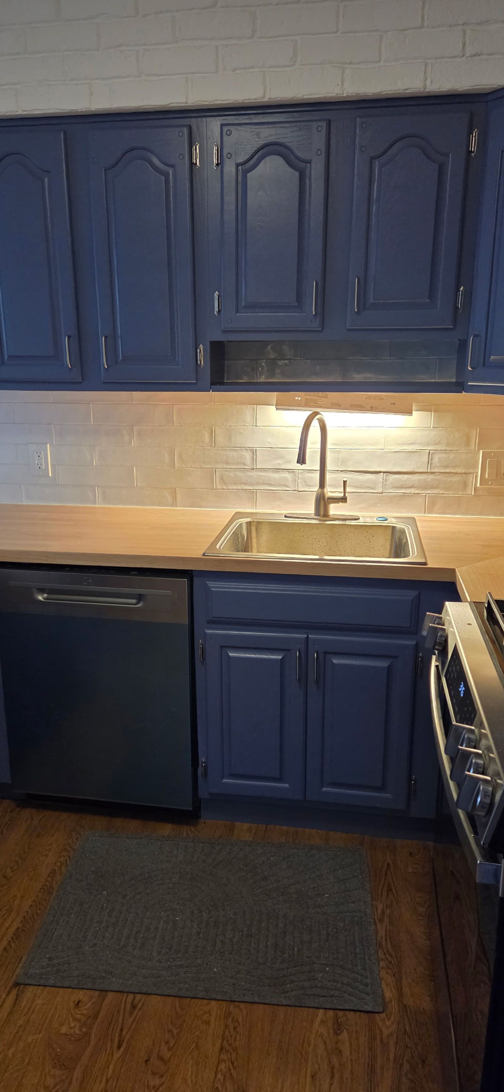 Kitchen with blue cabinets, a silver sink, and a wooden countertop. A dark rug is on the floor.