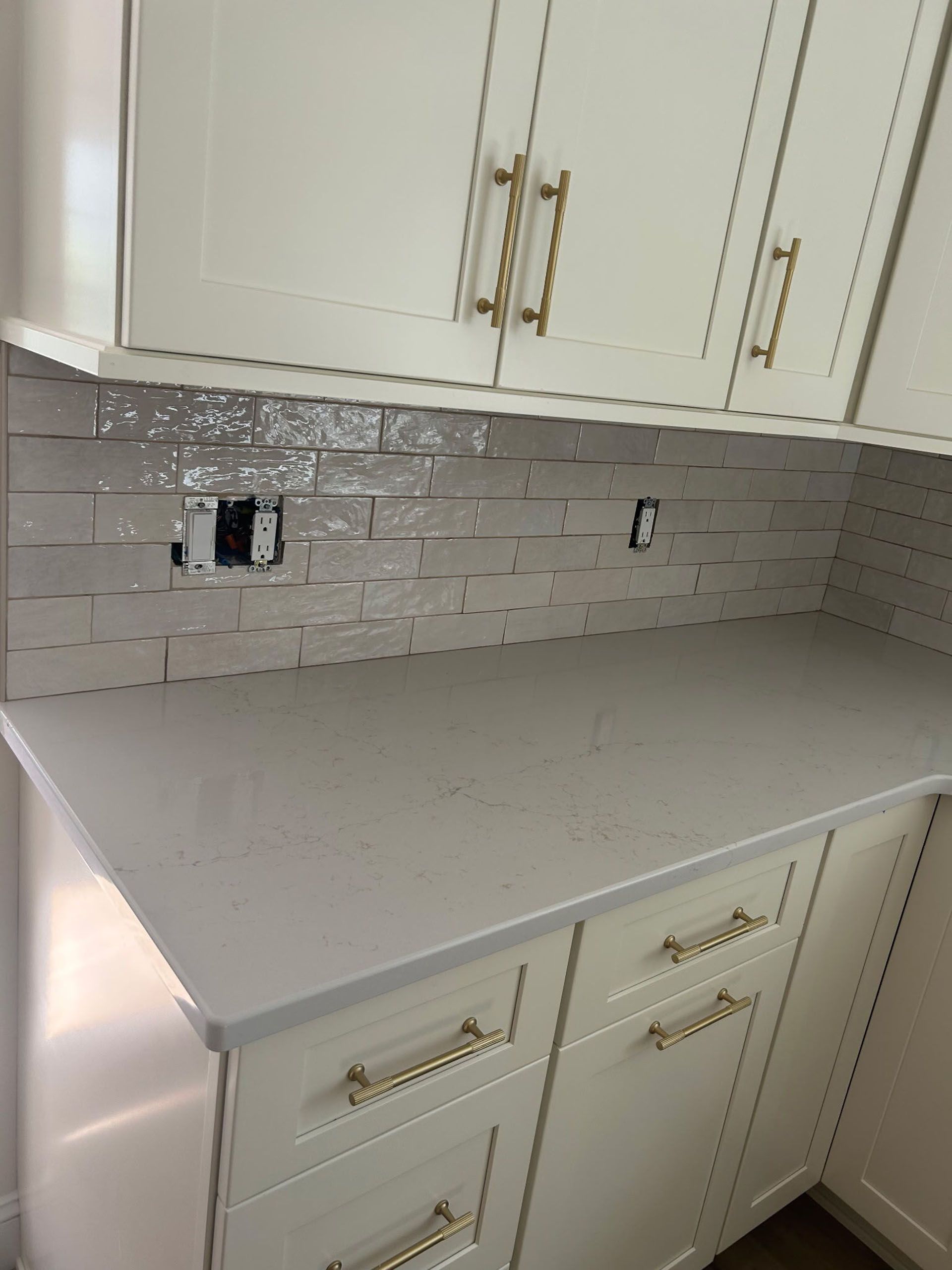 Cream-colored kitchen cabinets and countertop with gold hardware, white tile backsplash, and electrical outlets.