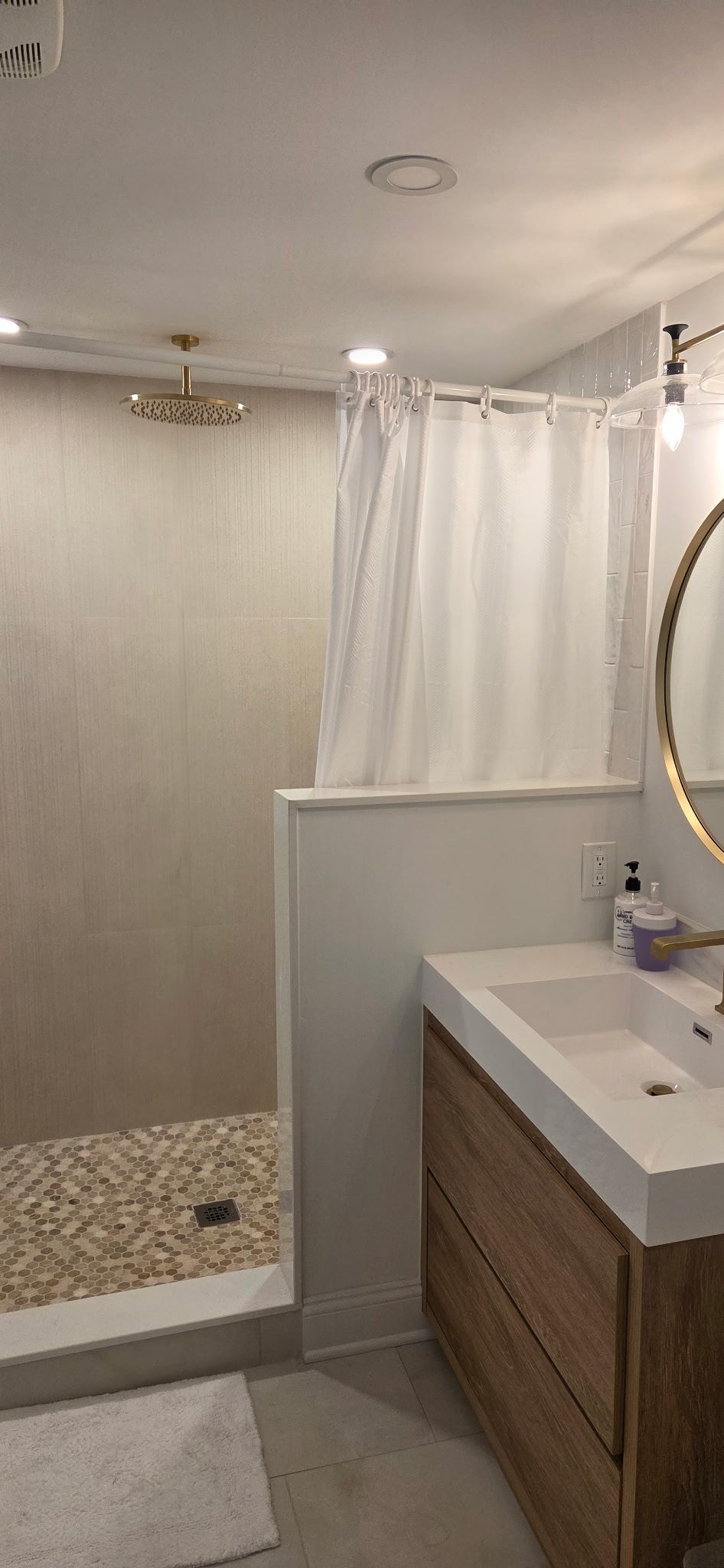 Bathroom with pebble-floor shower, wooden vanity, and gold-framed mirror. White shower curtain.