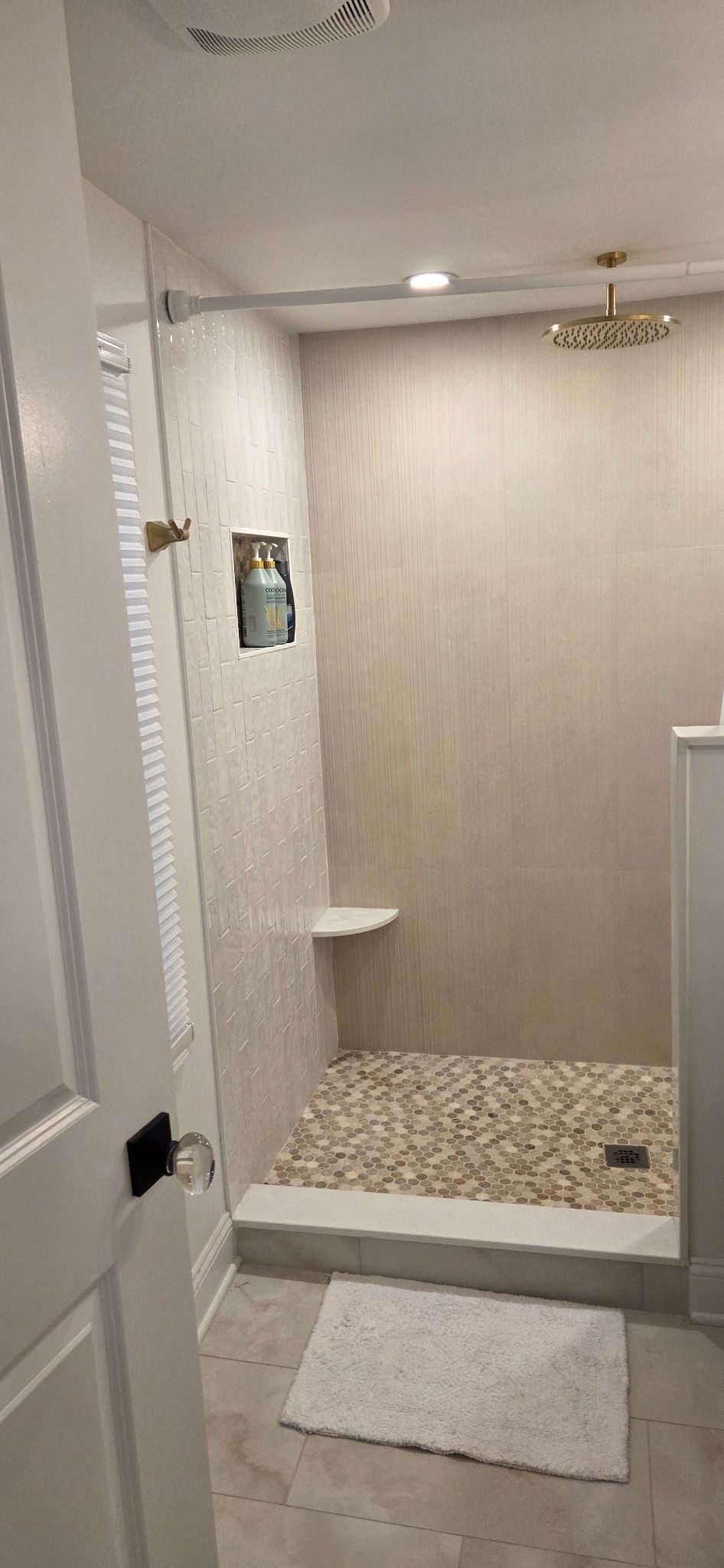 Bathroom with a pebble-floored shower, white walls, and a glass door. A white bath mat lies on the tiled floor.