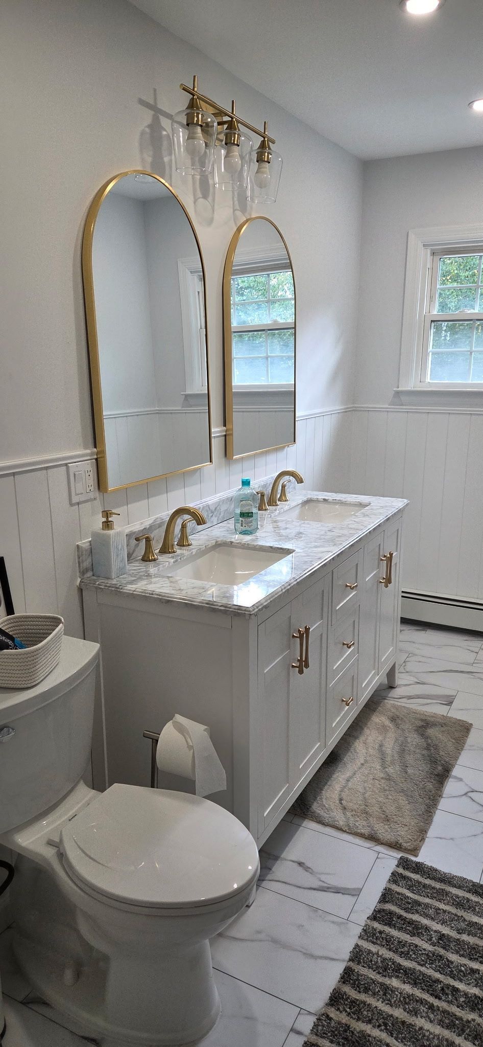 White bathroom with a double sink vanity, gold mirrors, and marble-look flooring.