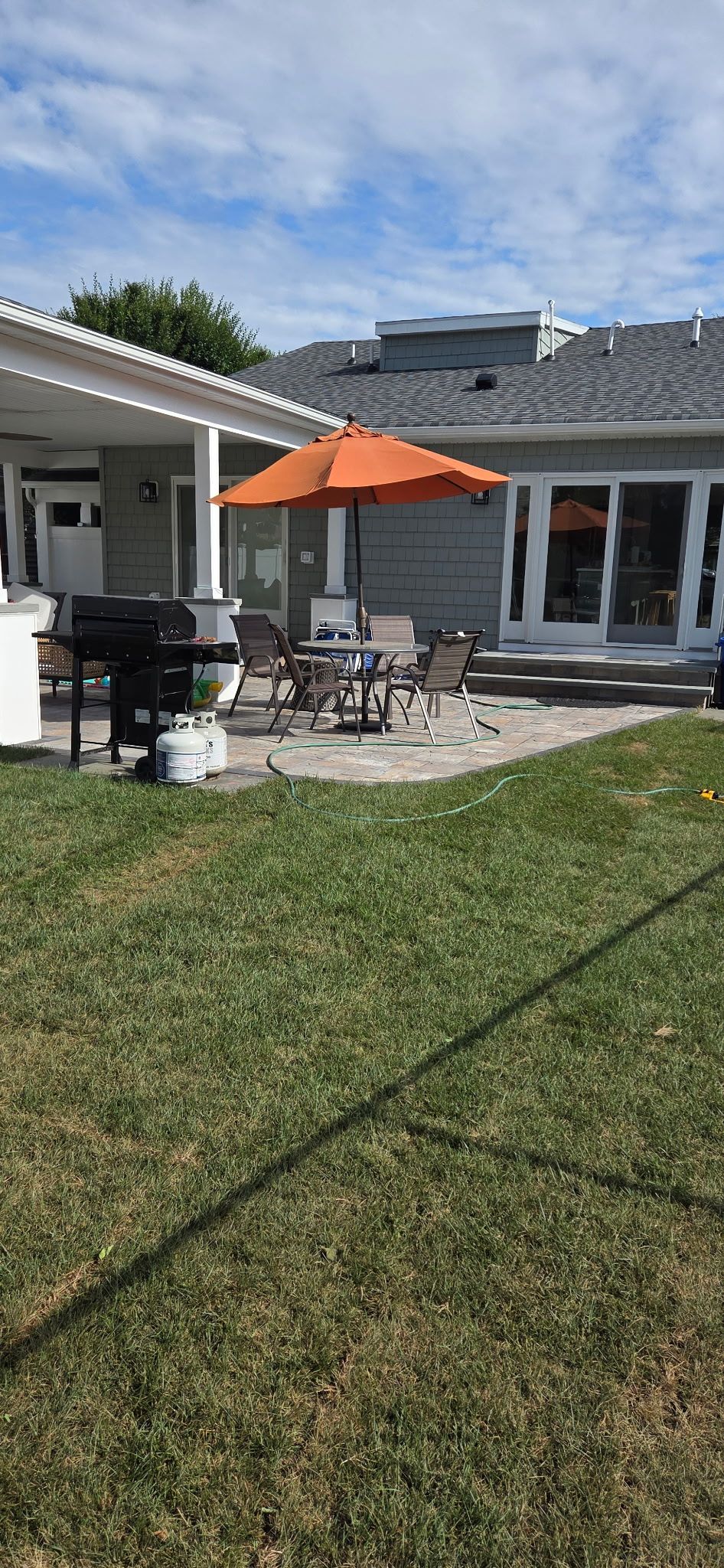 Backyard patio with orange umbrella, grill, and outdoor furniture on a sunny day.