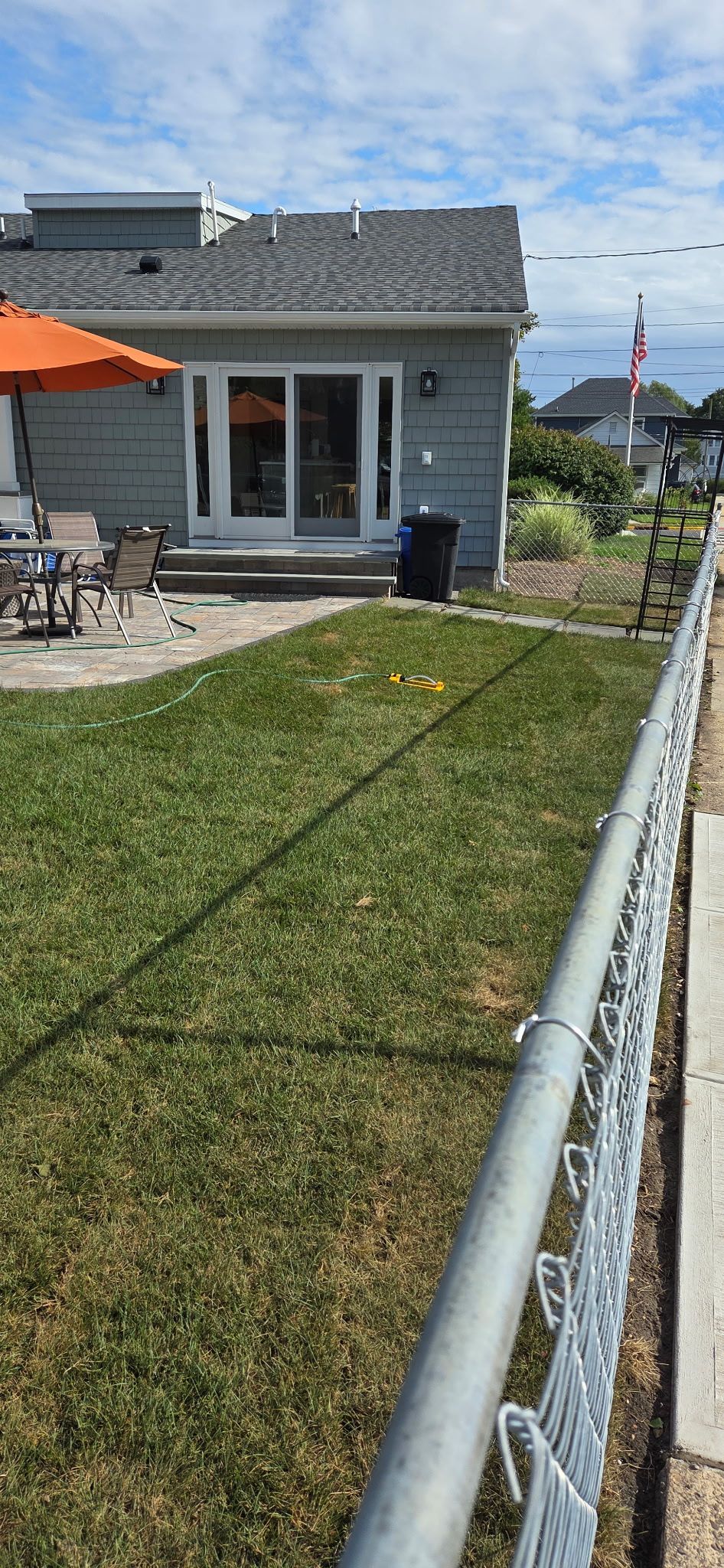 Backyard with gray house, patio with orange umbrella, and chain-link fence. Green grass and blue sky.