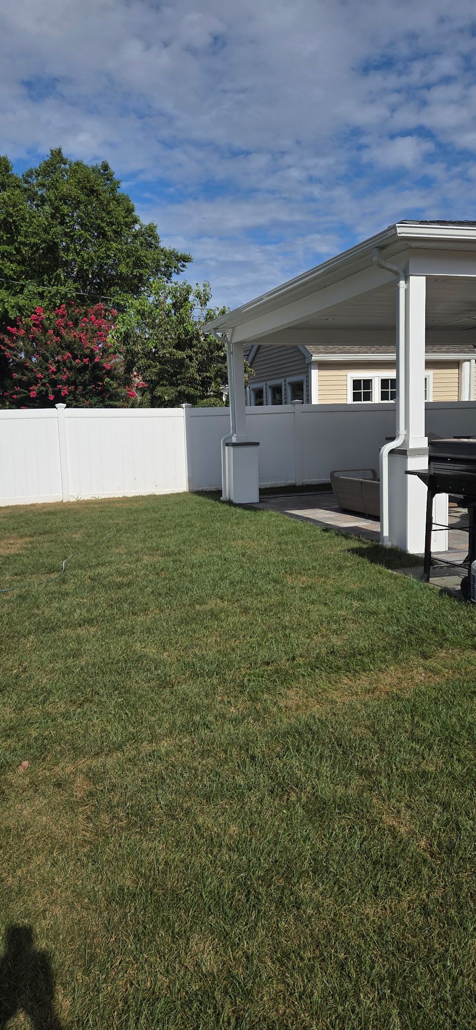 Backyard with green grass, white fence, and patio with a grill. Blue sky with clouds.
