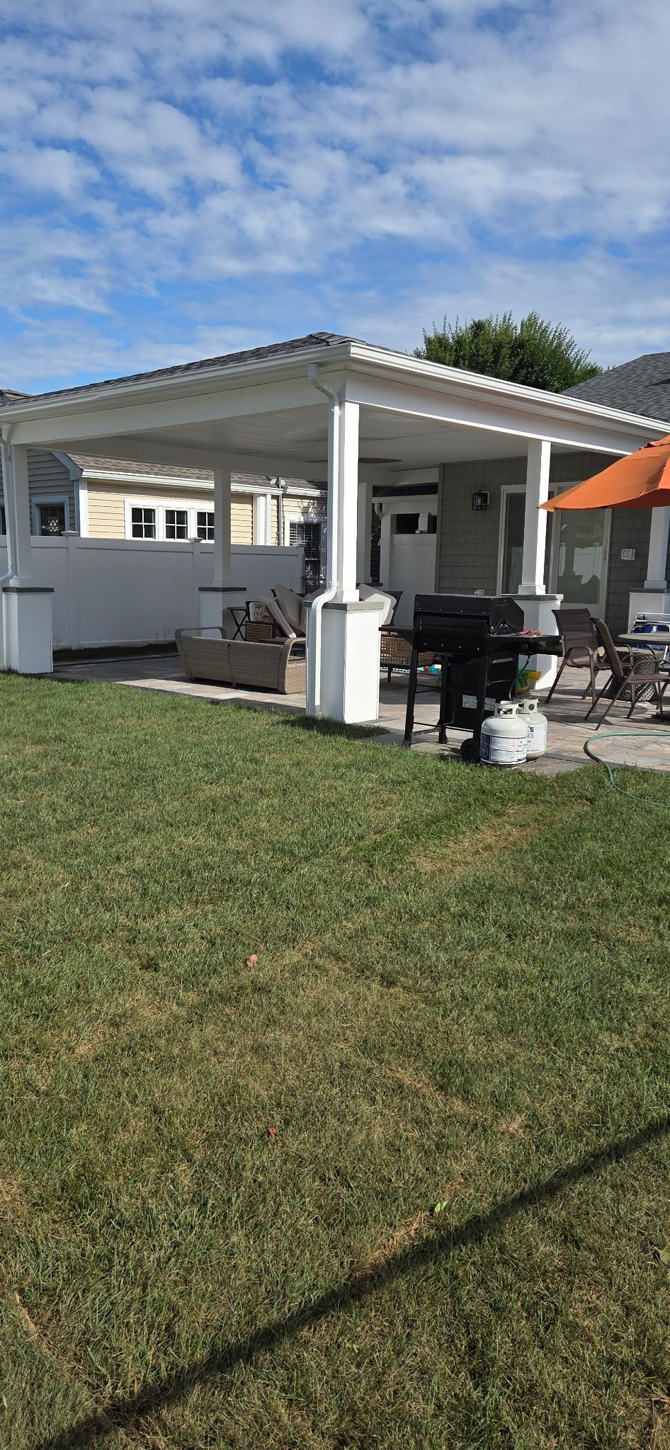 A white pergola covers a patio with a grill, outdoor furniture, and lush green lawn under a blue sky.