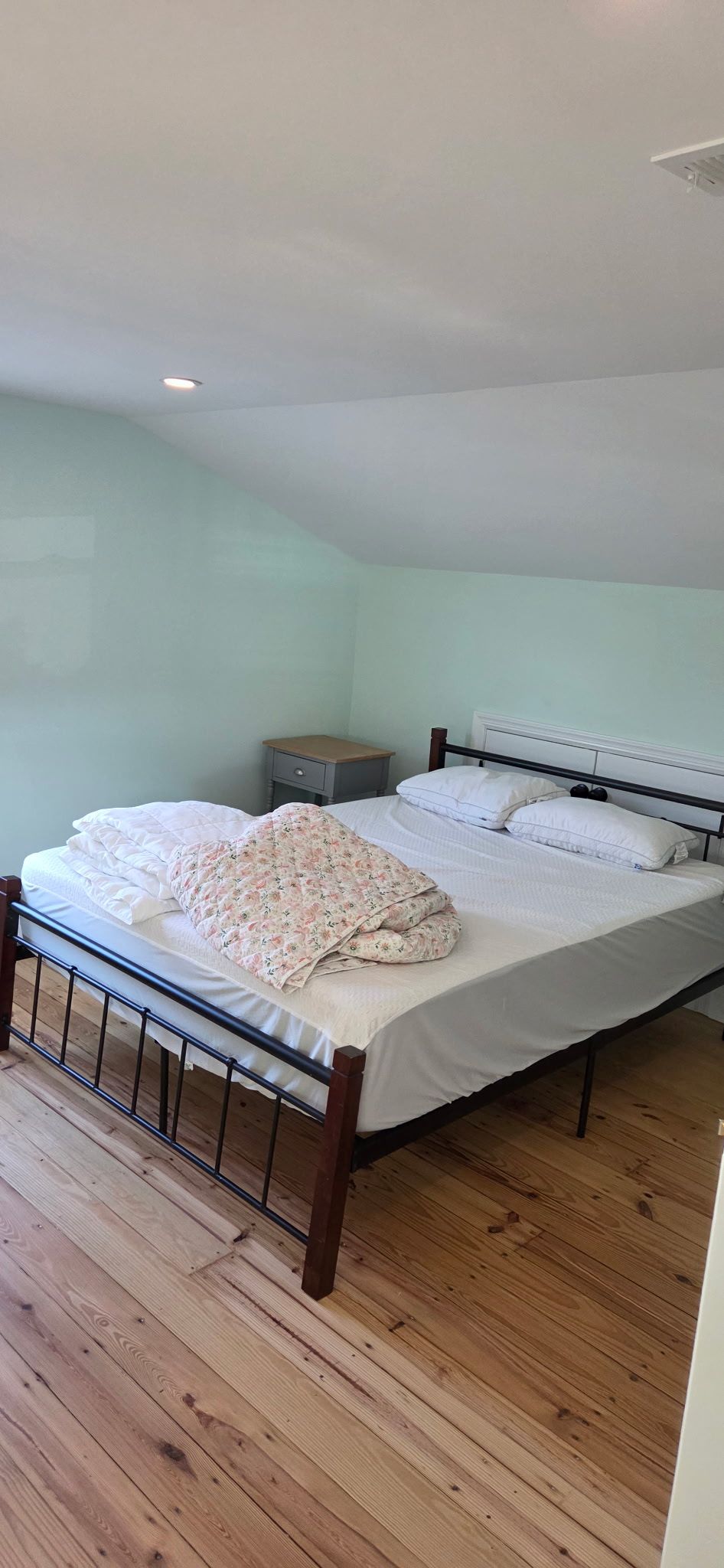 Bedroom with bed, pillows, patterned comforter, nightstand, and wooden floor. Light blue walls.