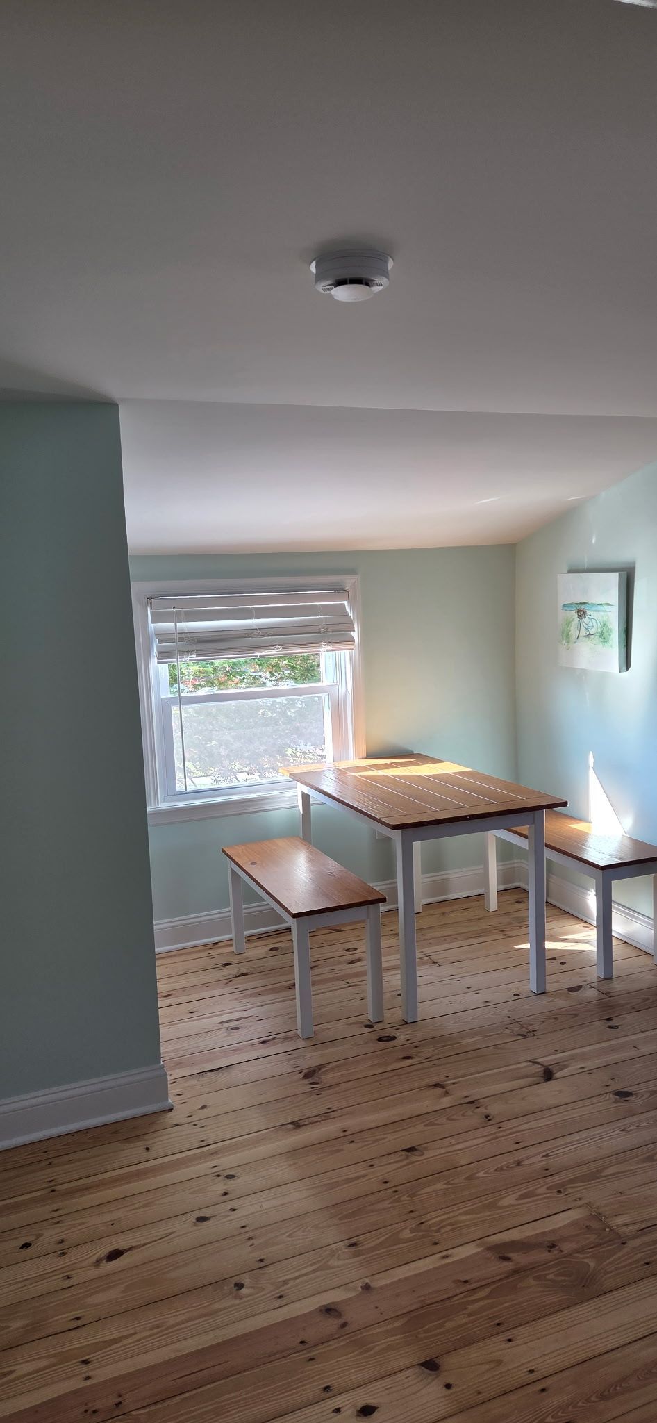 A dining room with wooden floors, a table, benches, and a window. Light blue walls and a white ceiling.