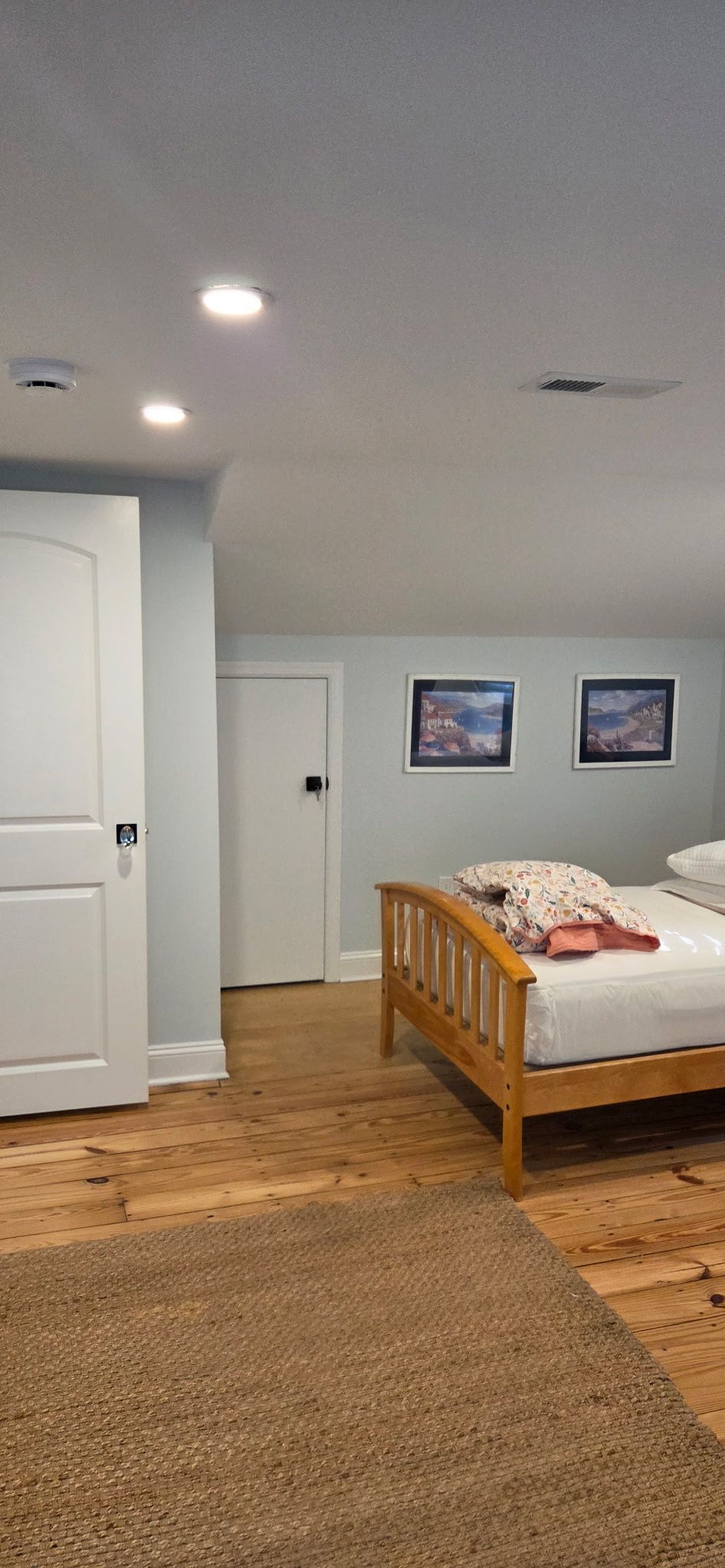 Bedroom with a bed, two doors, and framed pictures on the wall. The floor has a rug and wood planks.