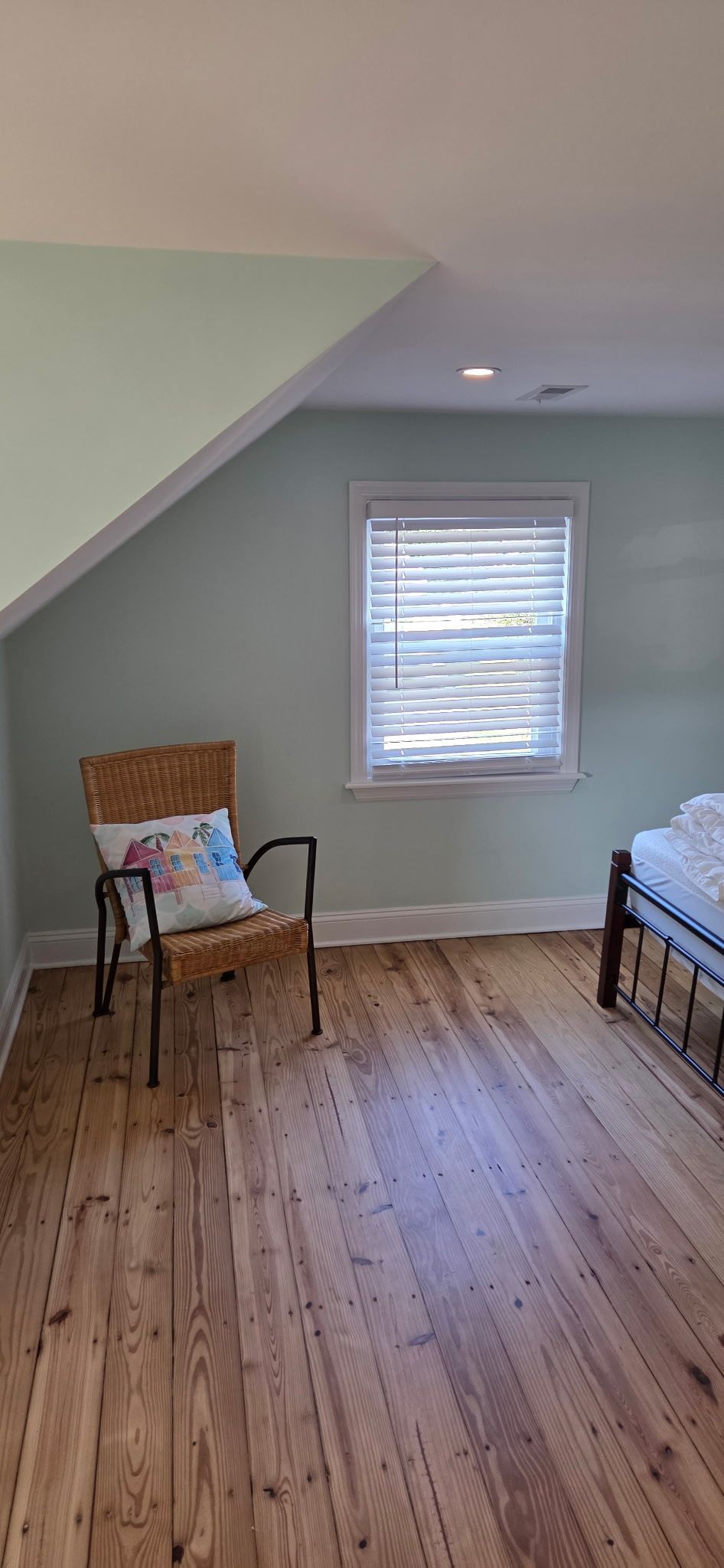 Bedroom with a woven chair, window, wooden floor, and a bed partially visible. Walls are light green.