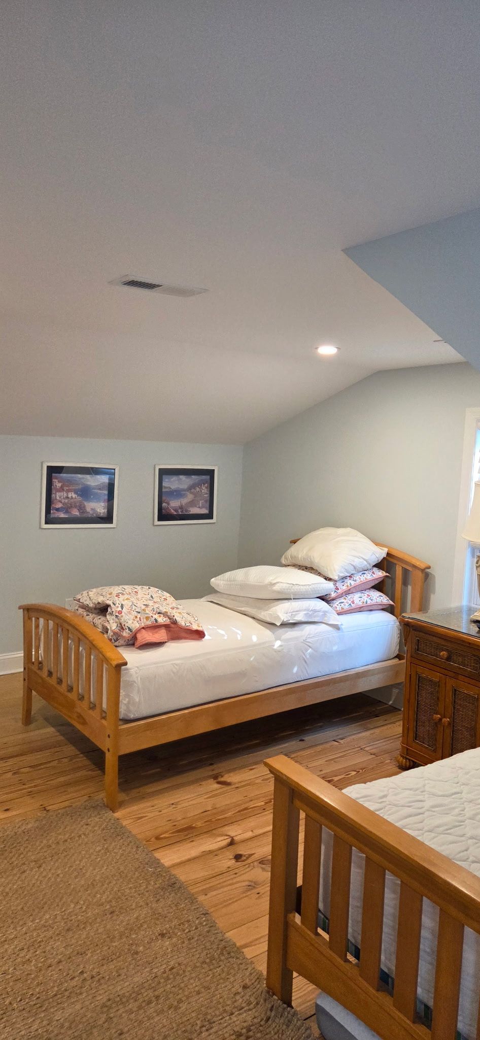 Bedroom with two beds, wooden frames, light blue walls, and framed art. Light wood flooring with a rug.