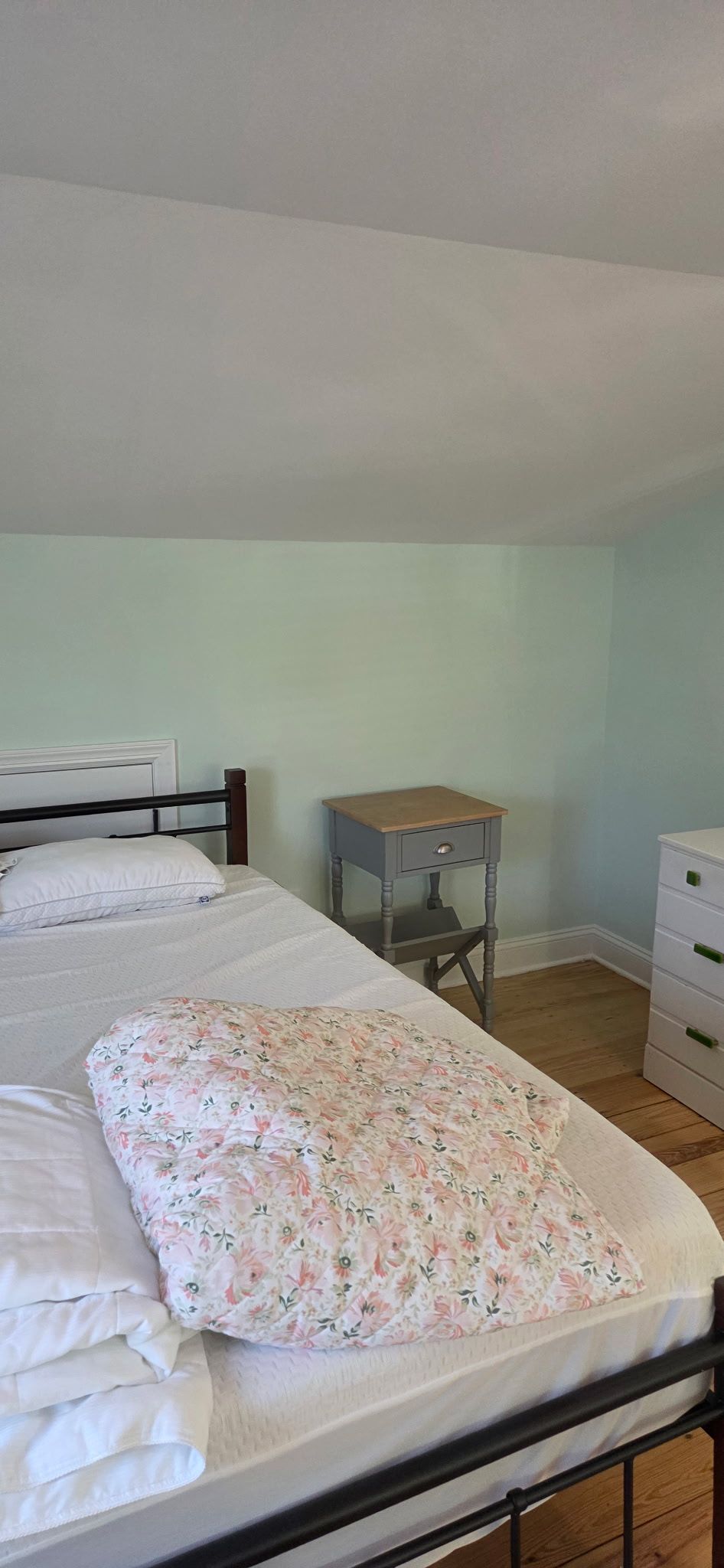 Bedroom with a bed, nightstand, dresser, and light green walls. The bed has a floral comforter and white sheets.