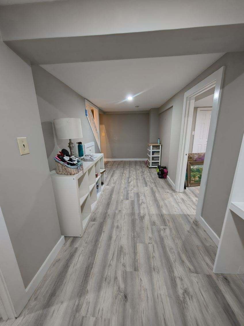 Narrow basement room with gray walls and wood-look flooring. White shelving and doorway are visible.